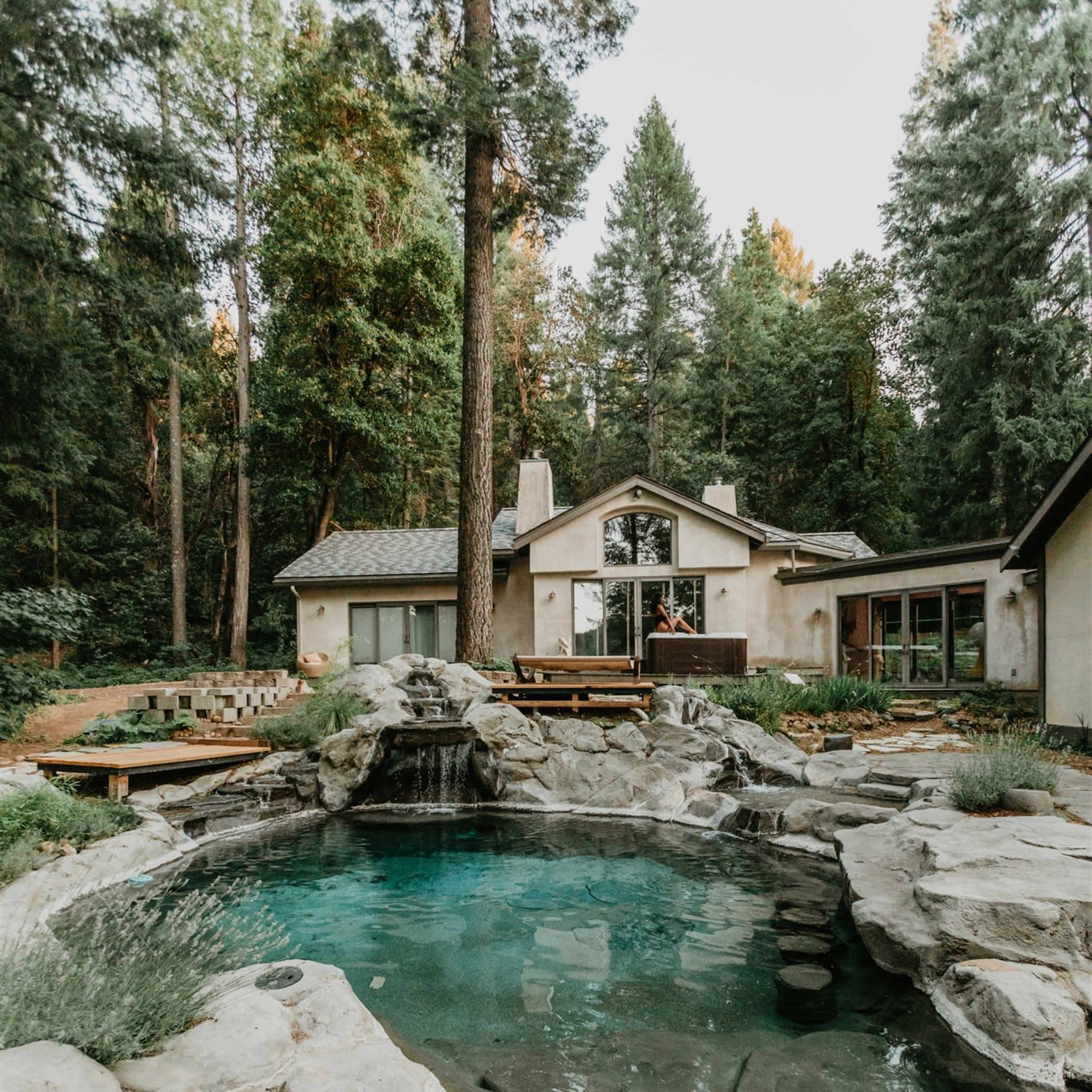 A rocky pool is surrounded by natural landscaping and a cabin-like house set among tall pine trees.
