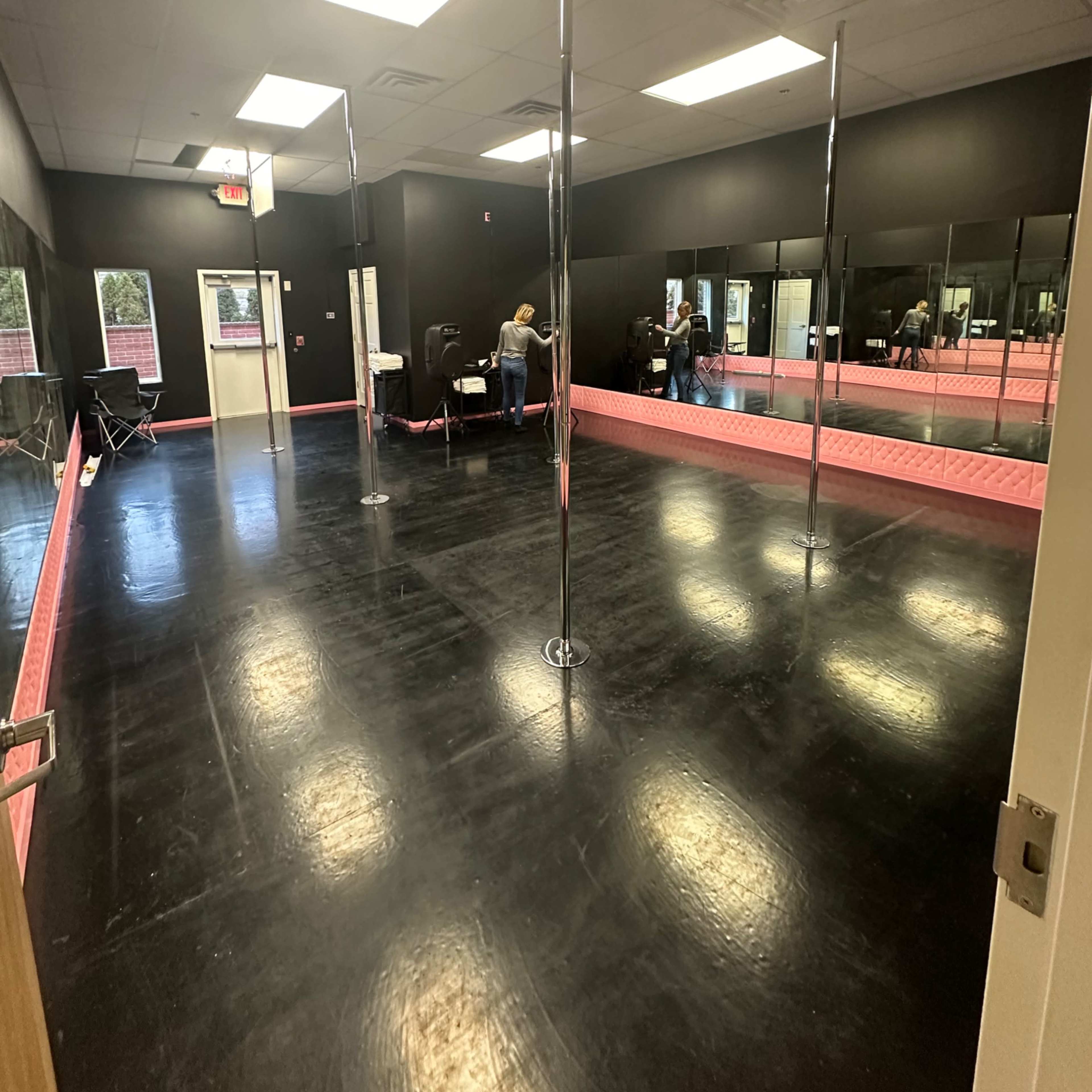 A dance studio with black walls, a polished floor, and several poles arranged in an open space, featuring mirrors along one side.