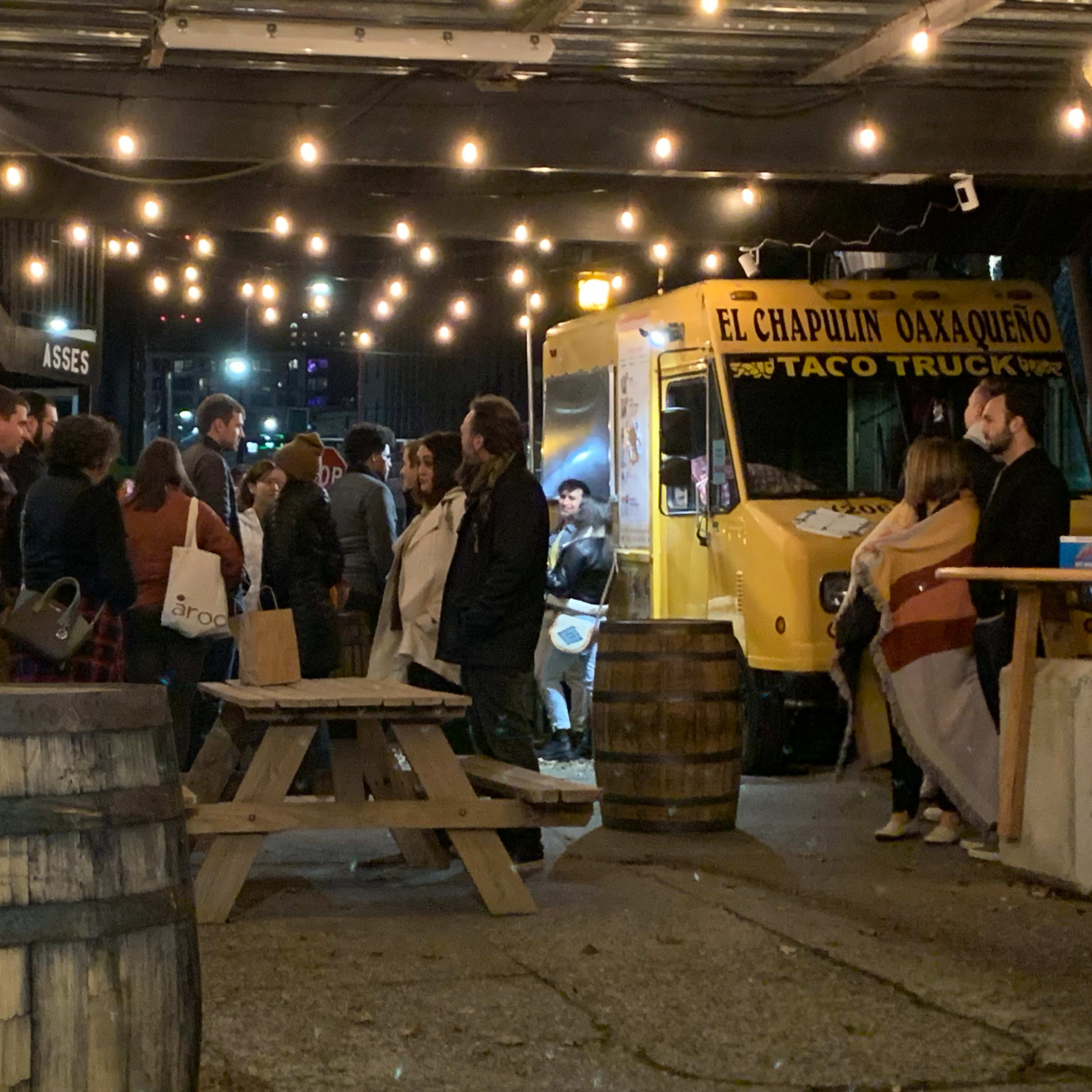 A group of people gathers around a yellow taco truck under string lights in a courtyard with wooden tables and barrels.
