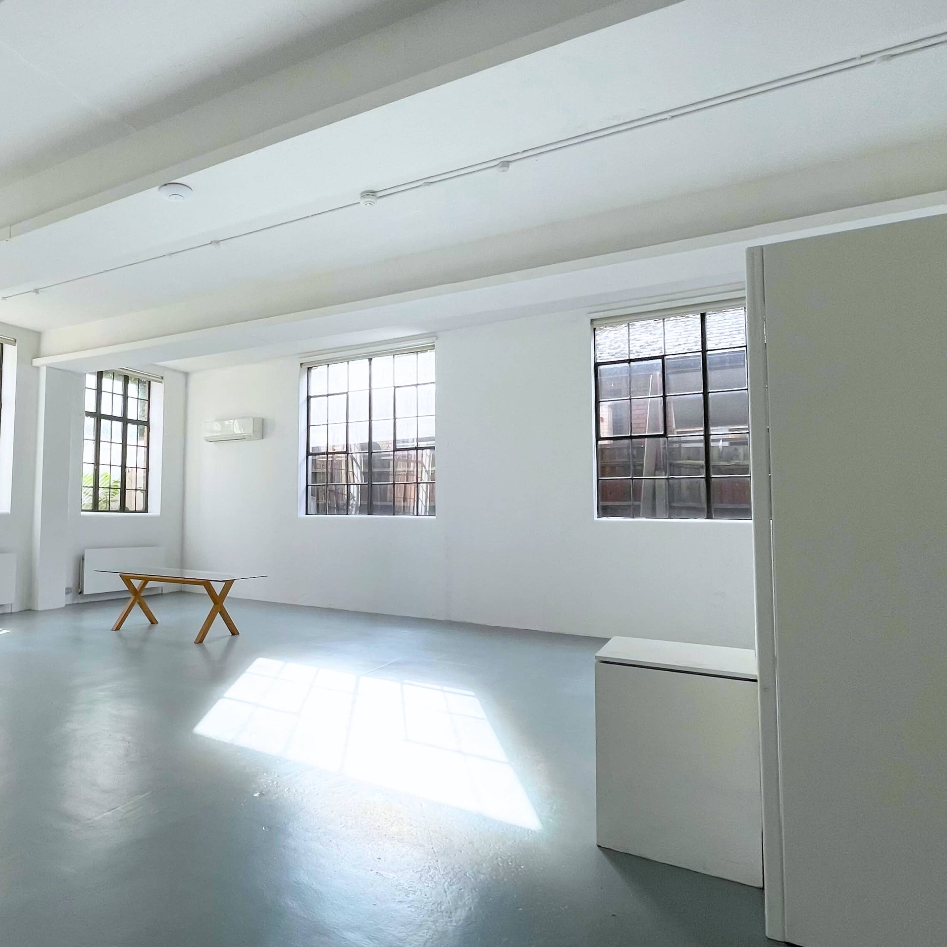 A spacious, well-lit room with large windows, a wooden table in the center, and minimal furnishings.