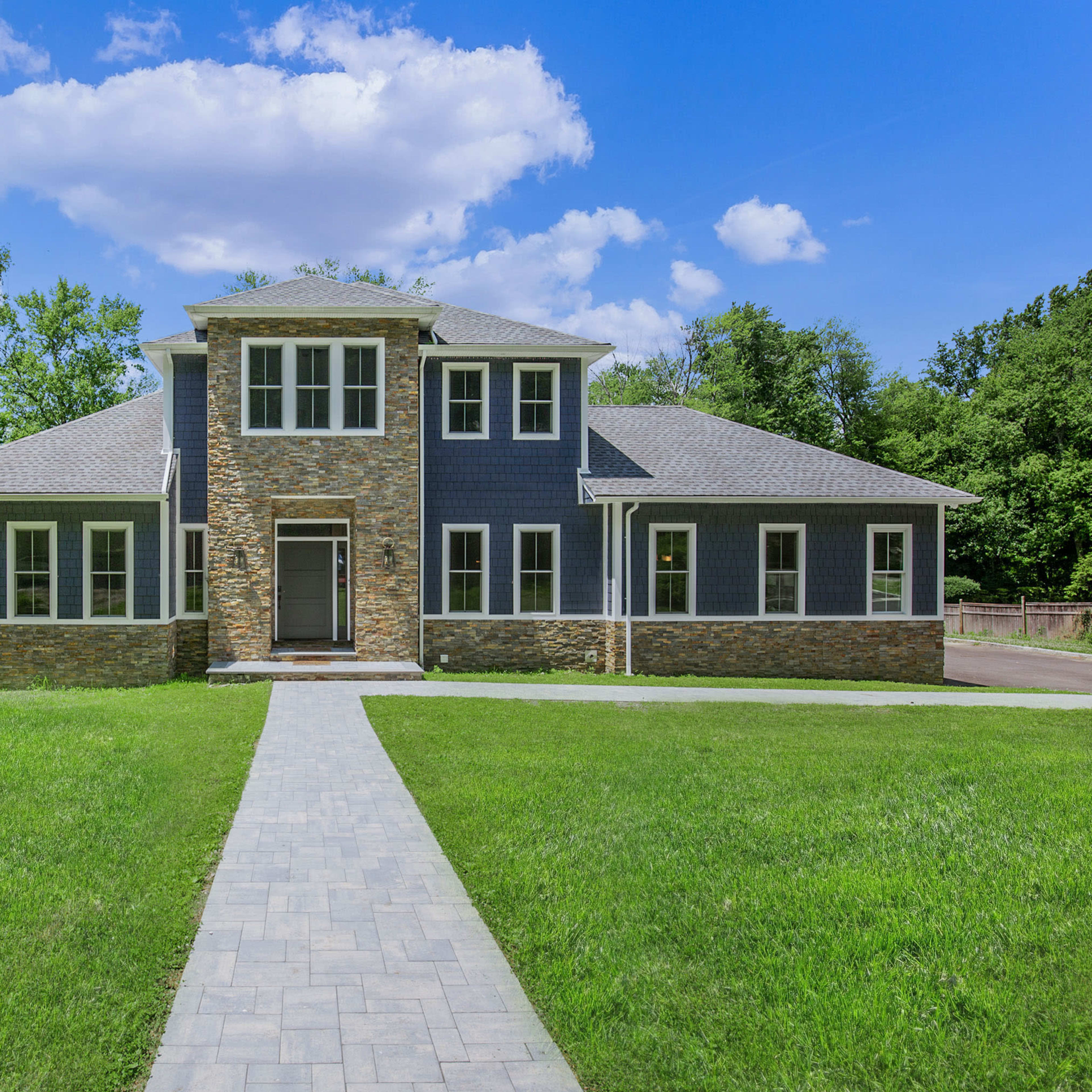 A large, modern two-story house with a stone and wood exterior, surrounded by a grassy area and trees under a blue sky.