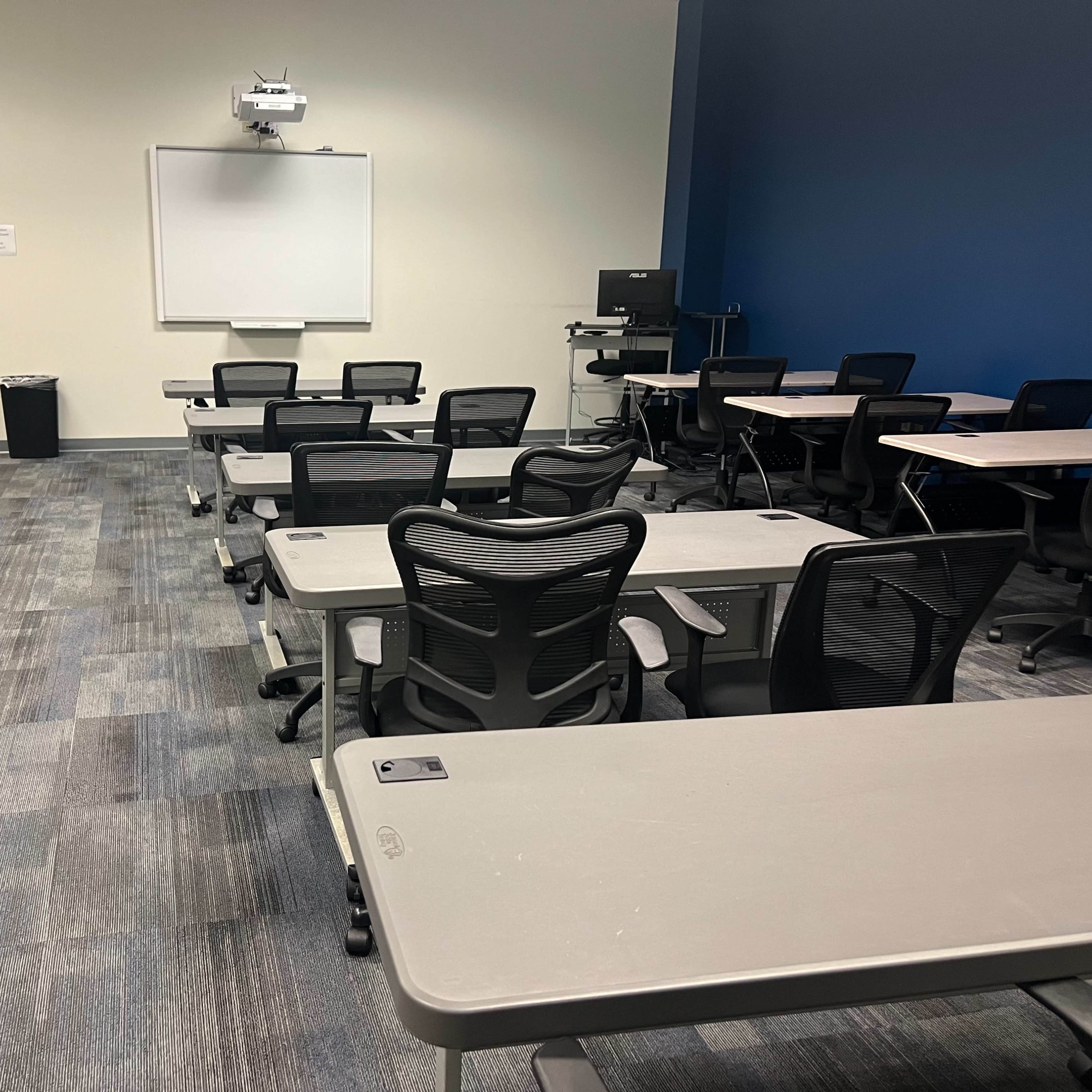 A classroom setup with several rows of desks and chairs, a whiteboard, and a projector mounted on the ceiling.