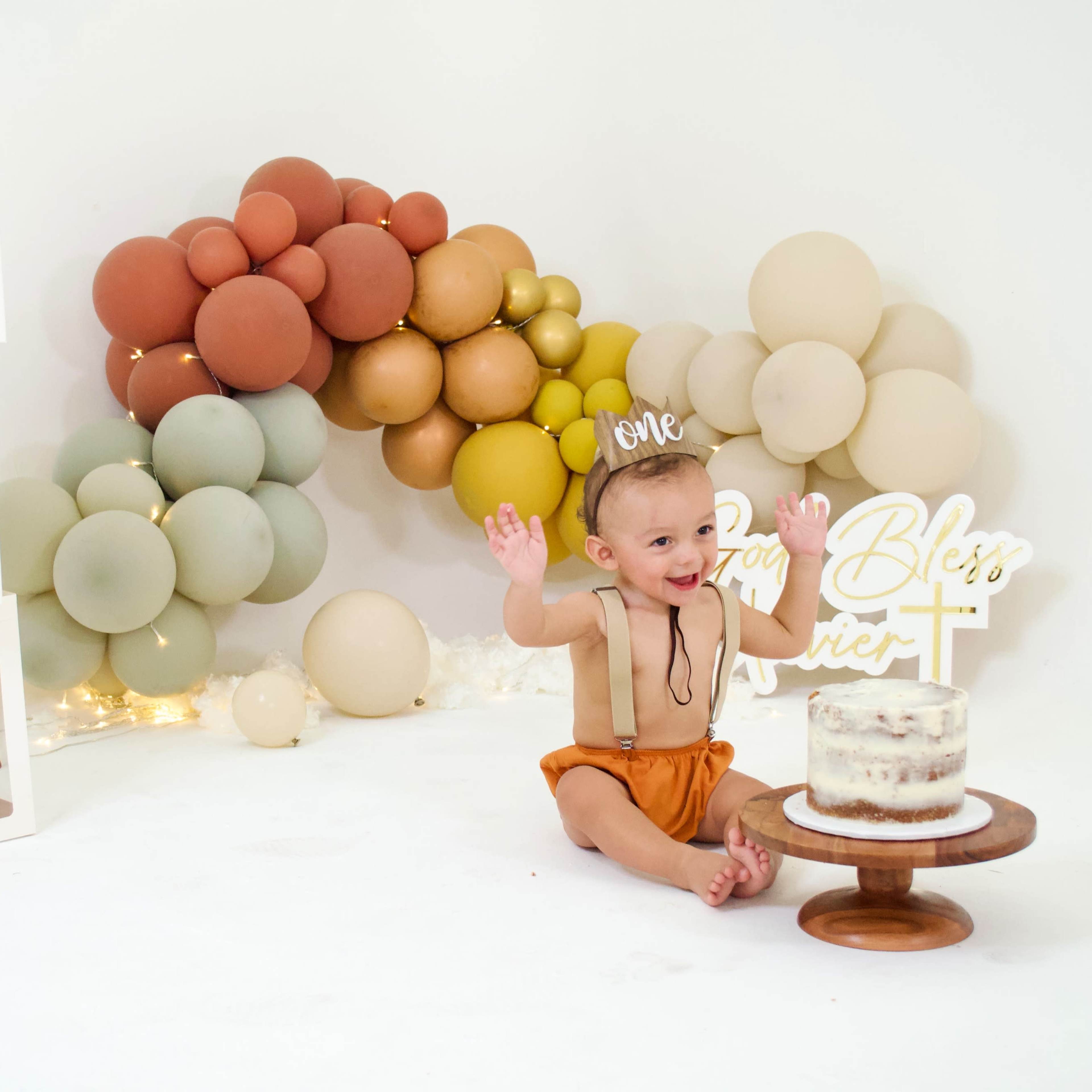A baby wearing a party hat sits beside a decorated cake on a wooden stand, with balloon arrangements and a sign that reads "God Bless" in the background.