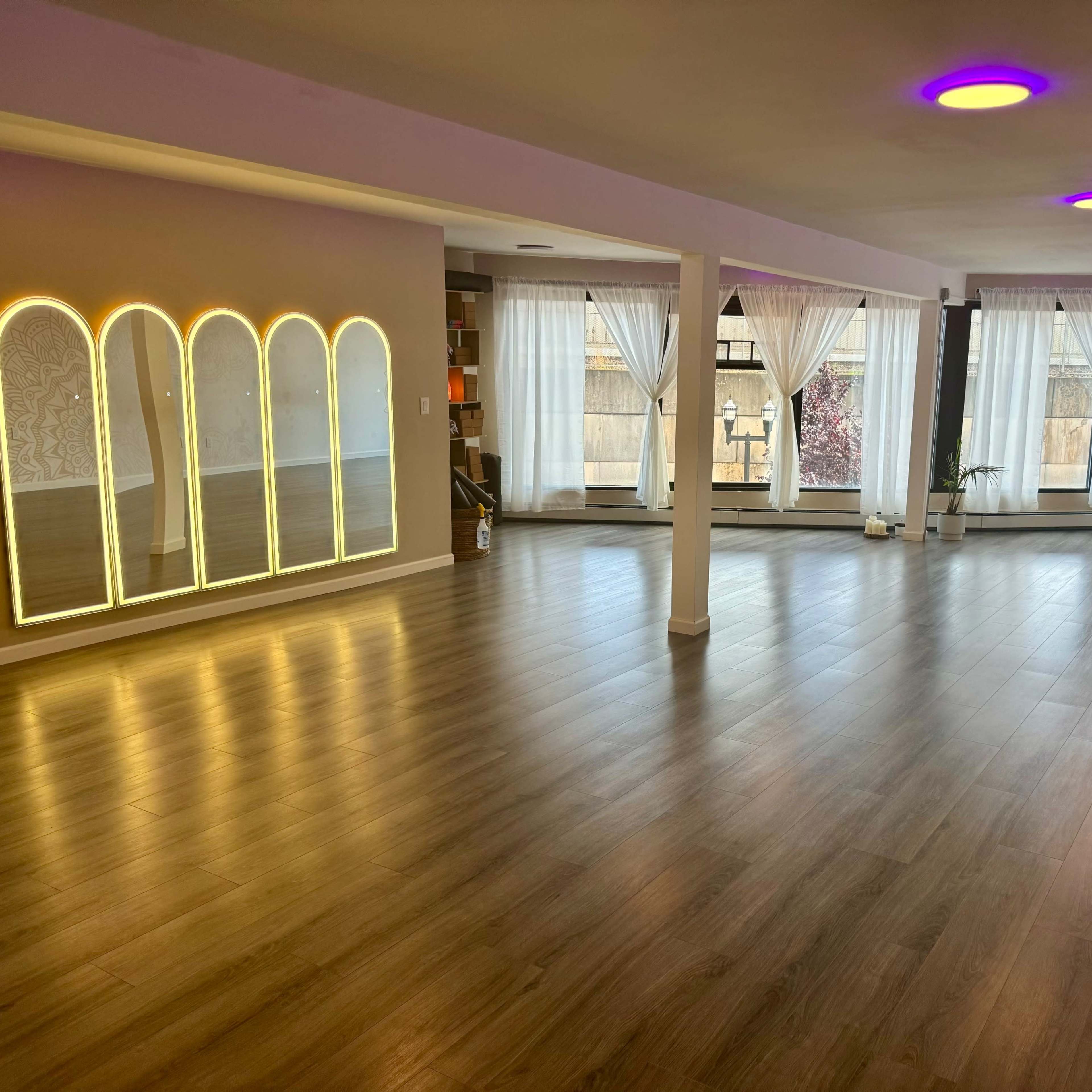 An empty room with wooden flooring, large windows covered with sheer curtains, and a decorative mirror with illuminated arches along one wall.