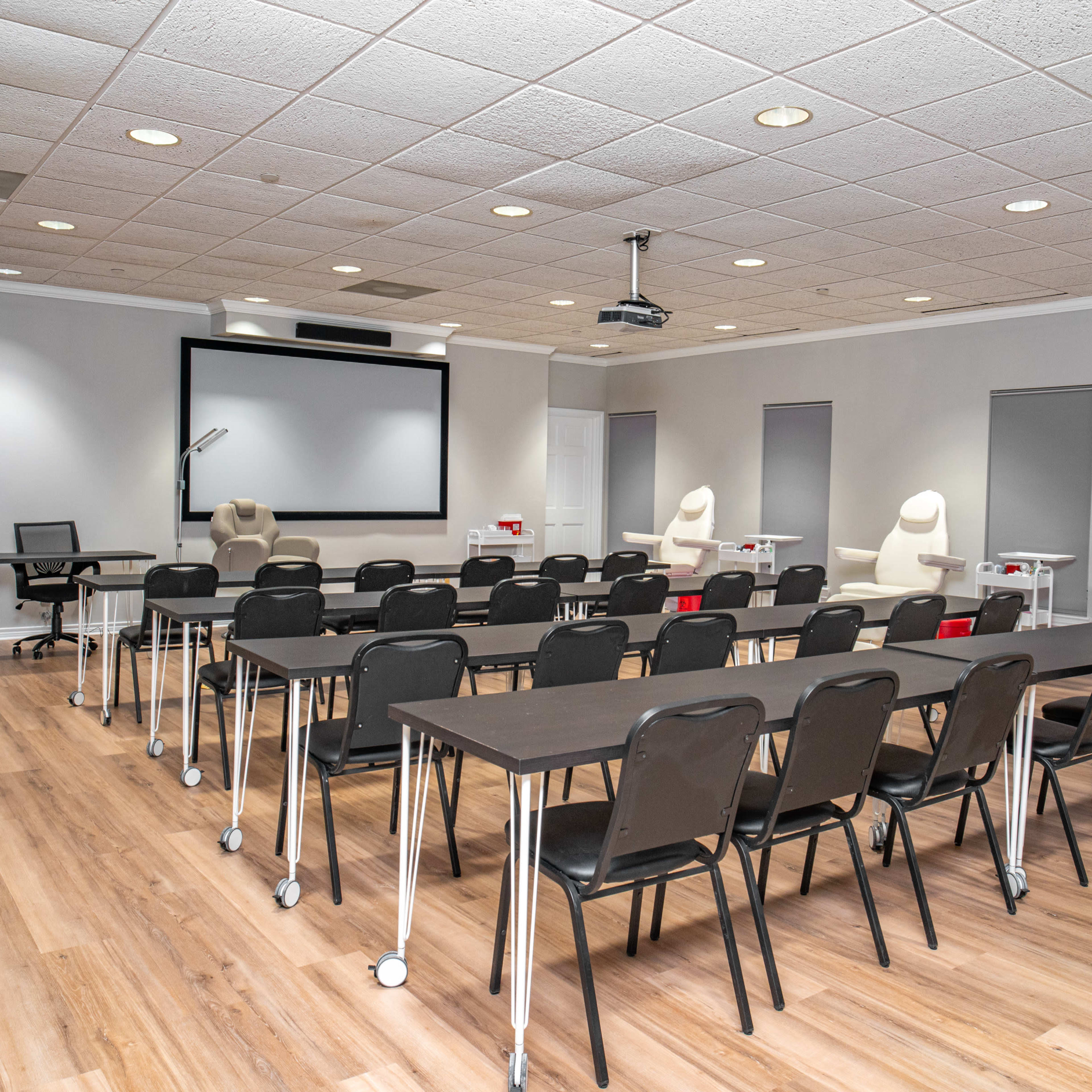 A training room features rows of black chairs and long tables facing a projection screen, with additional equipment and supplies in the background.