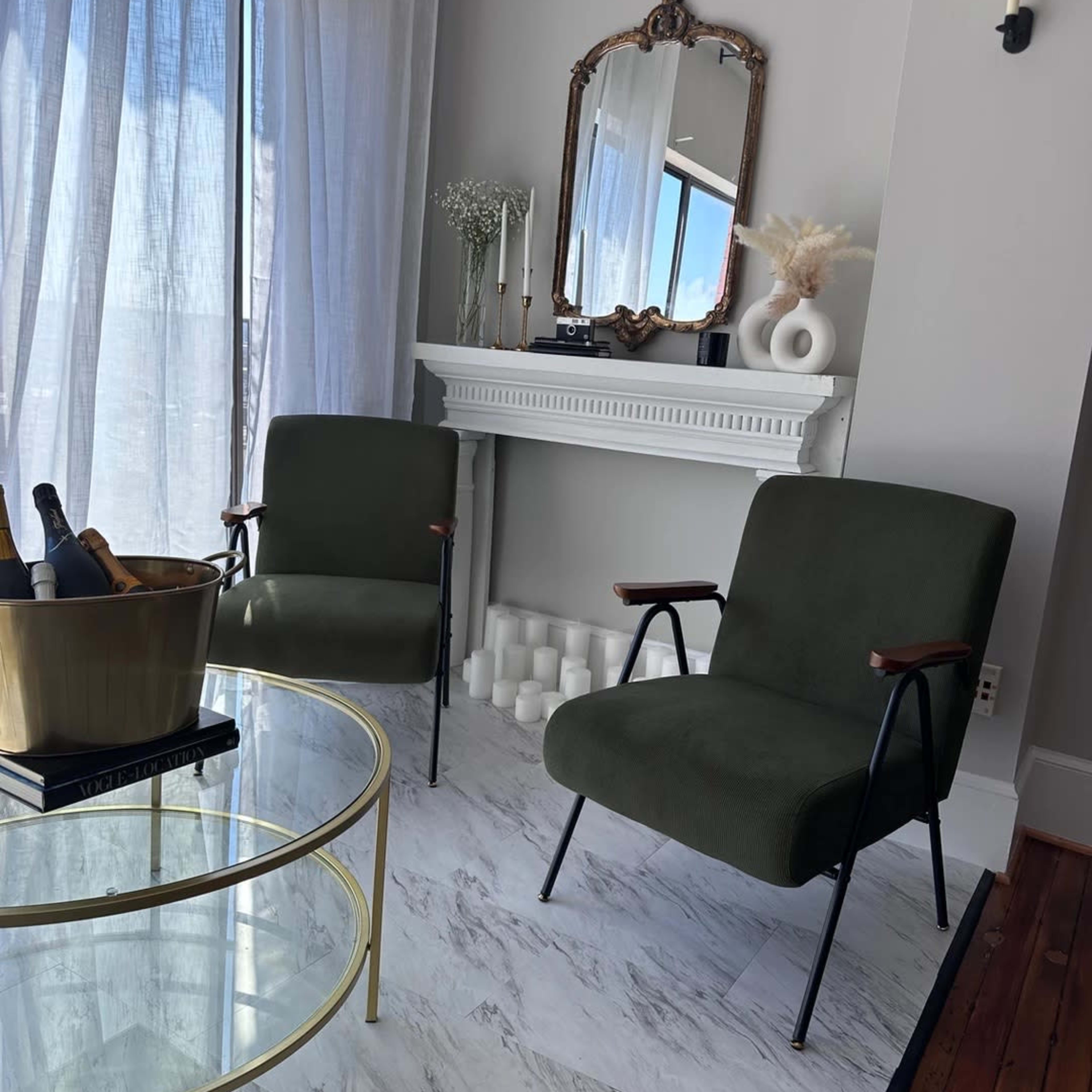 A modern living room with two green chairs facing a glass coffee table, near a fireplace with decorative items and soft natural light coming through sheer curtains.