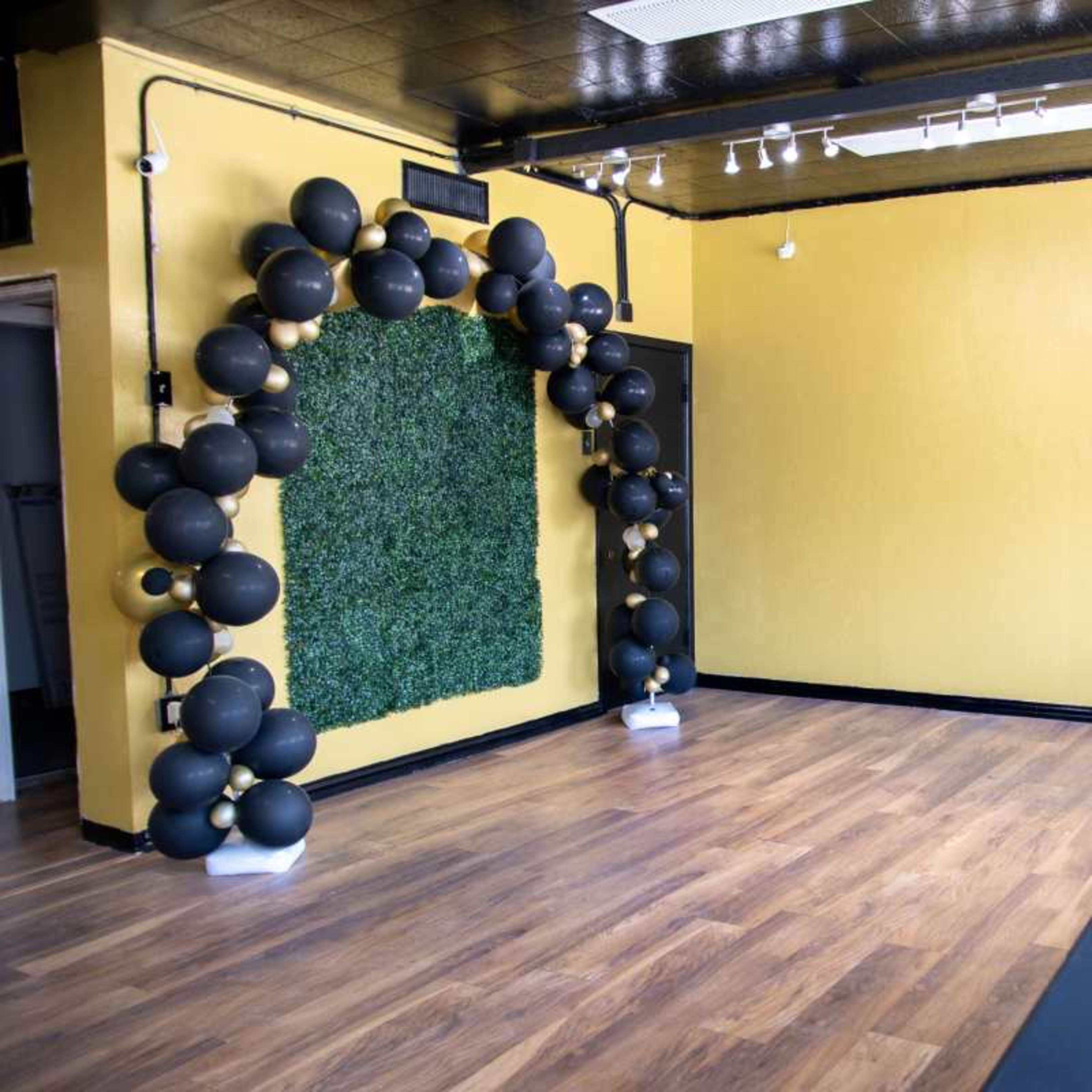 An empty room with yellow walls, wooden flooring, and a decorative arch made of black balloons next to a wall covered in green foliage.