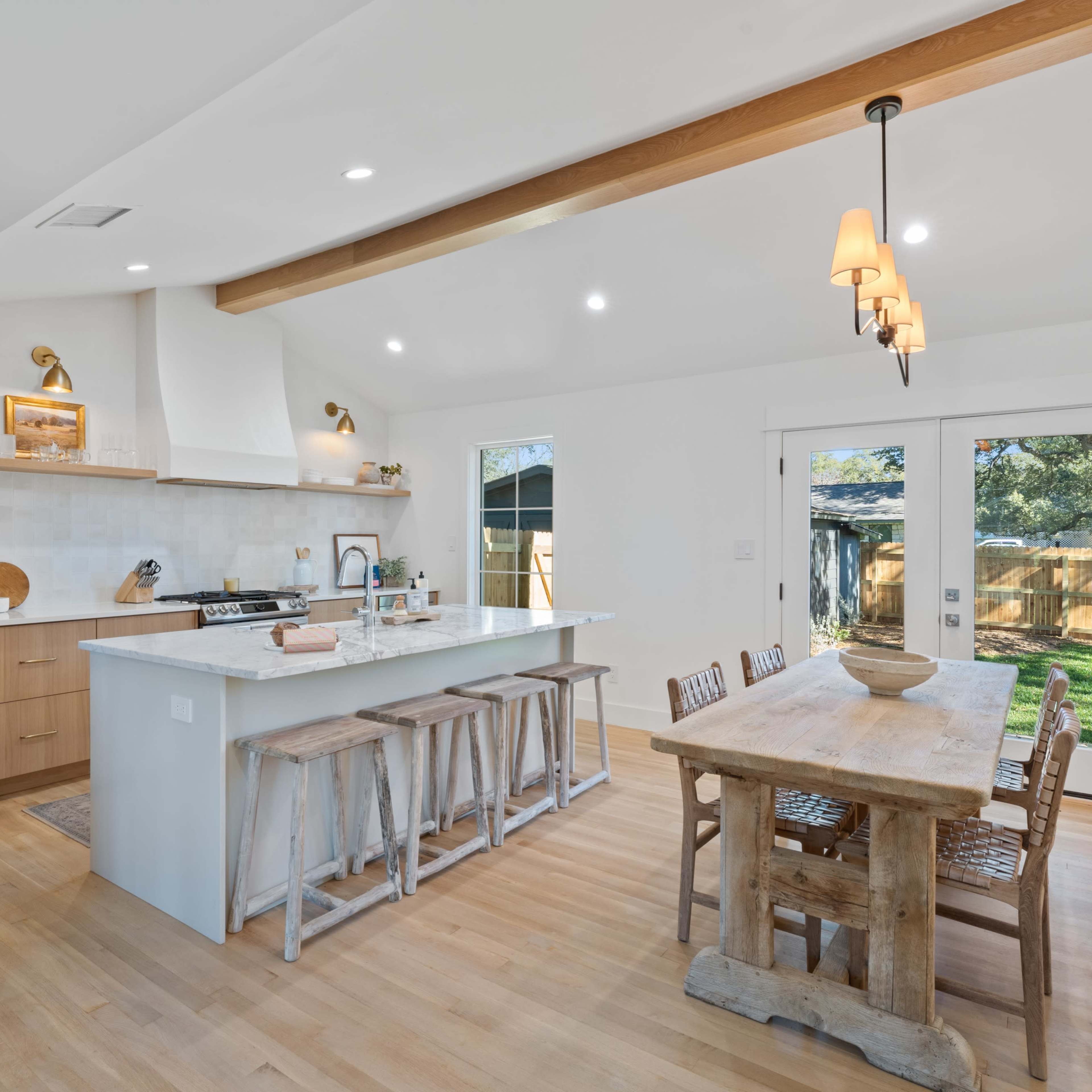 A modern kitchen features a large island with seating, wooden beams on the ceiling, and a rustic dining table under pendant lights.