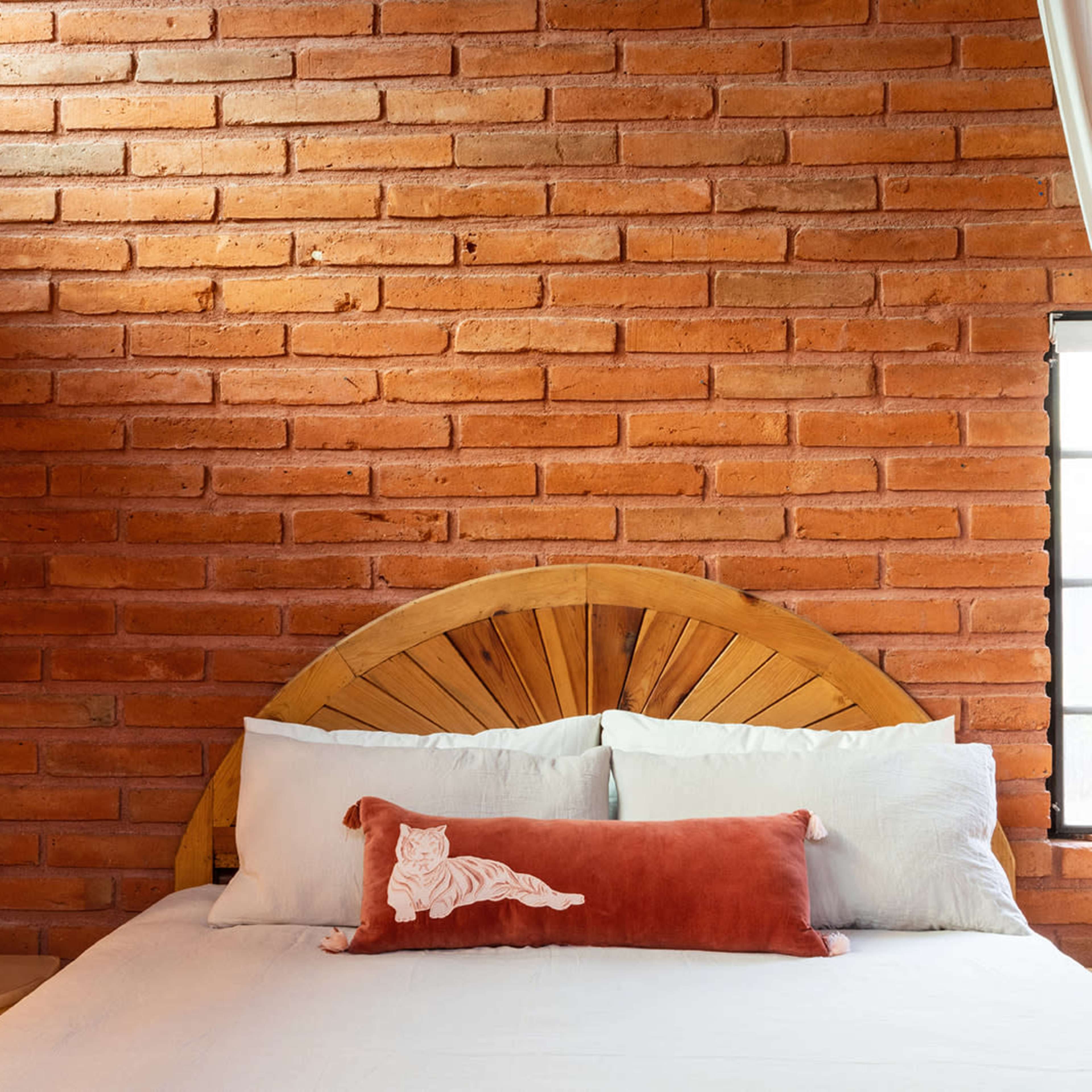 A bed with a wooden headboard and decorative pillow is positioned against a brick wall, complemented by a window and lamps on either side.