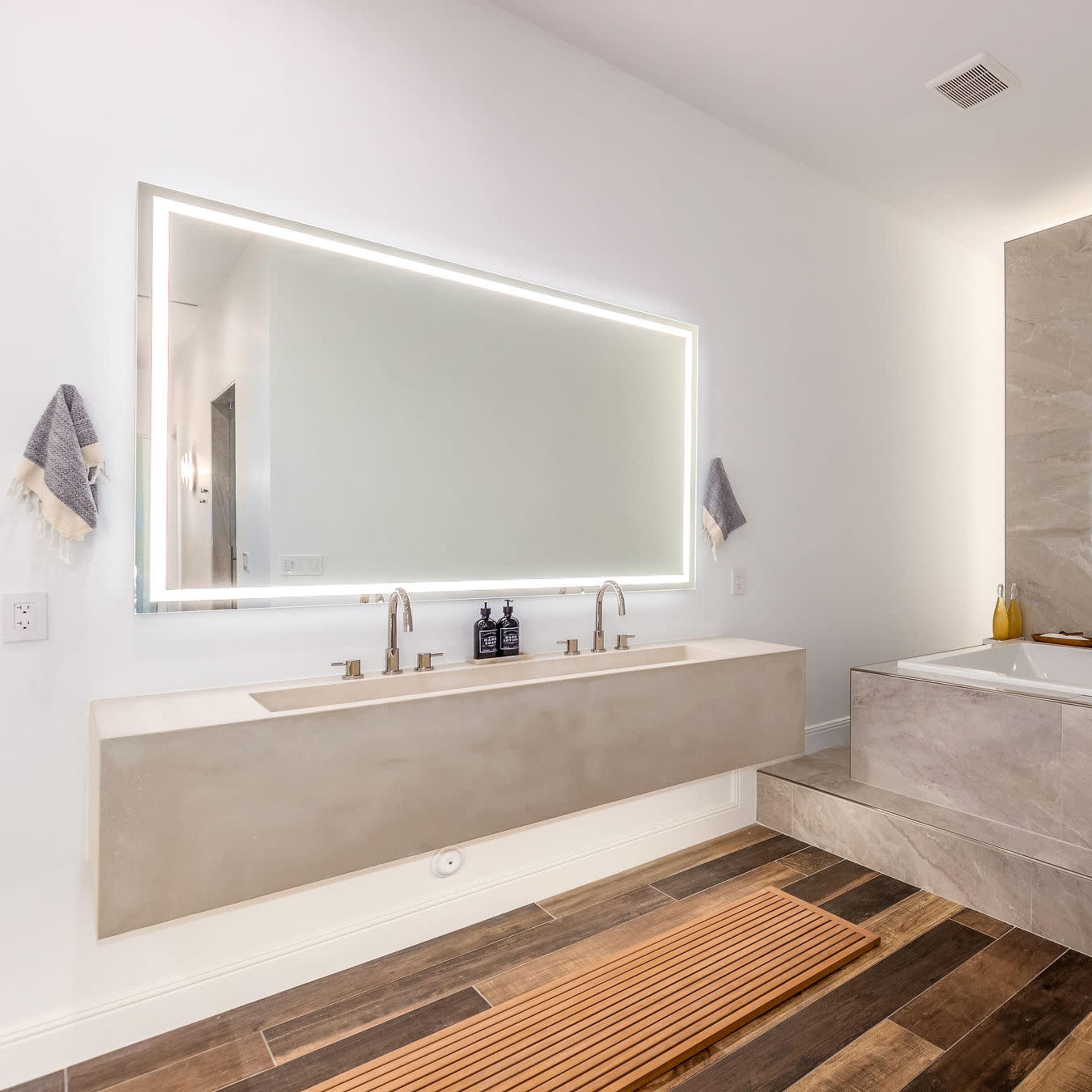 The bathroom features a large mirror with integrated lighting, a dual sink vanity made of concrete, a freestanding bathtub adjacent to a marble wall, and wood flooring.