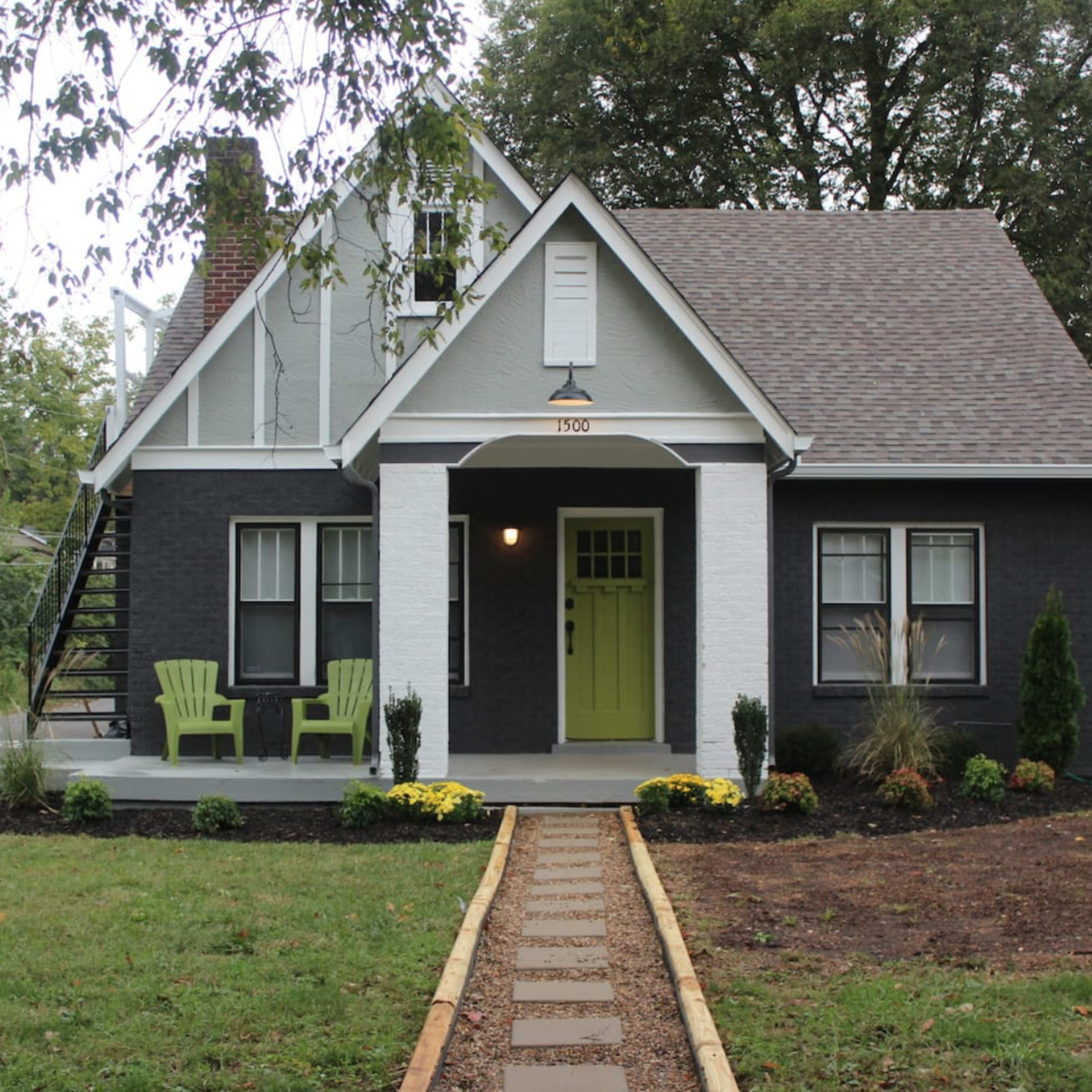 A two-story house with dark siding and a white trim features a green front door and a pathway lined with flowers and trees.