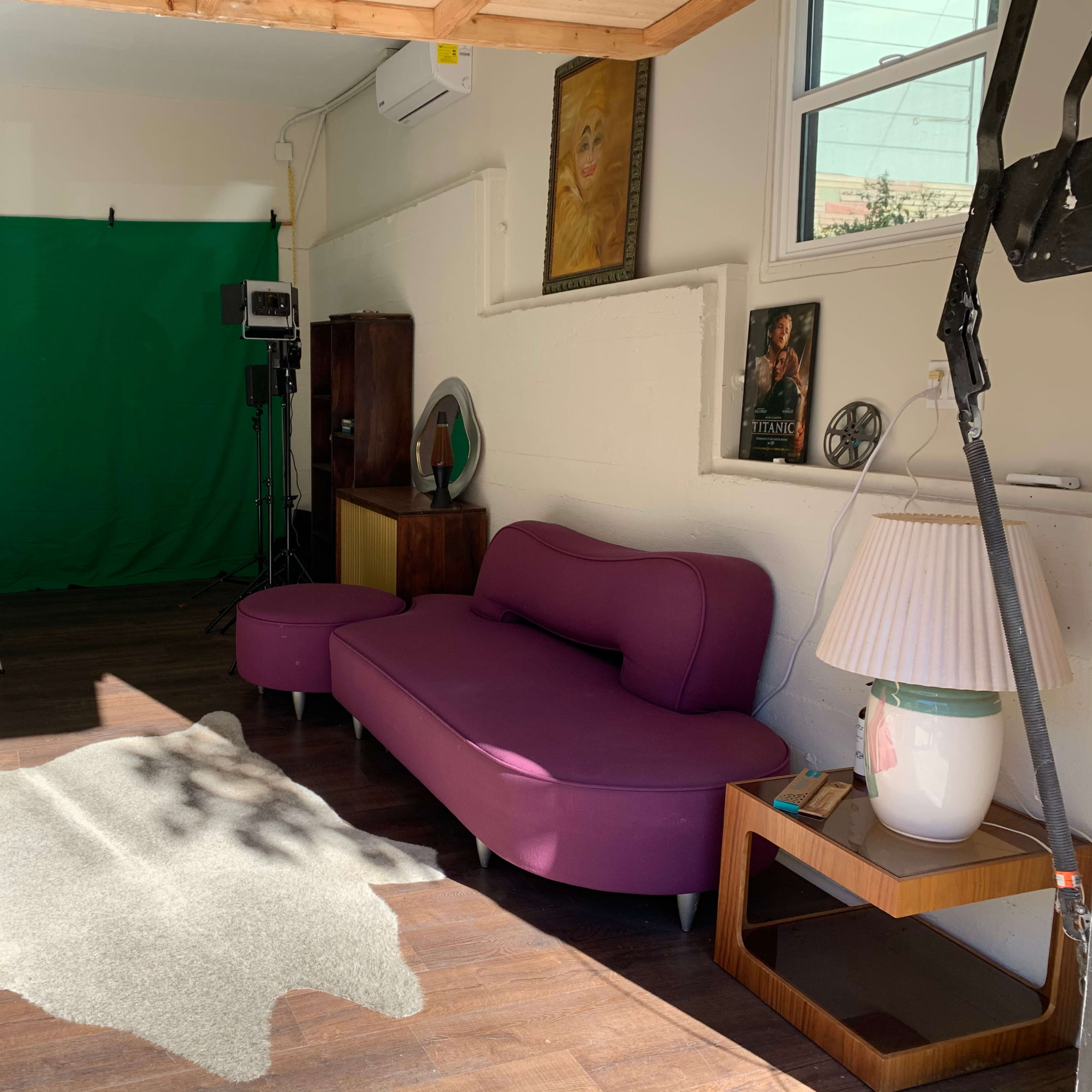 A studio space features a green backdrop, two lights, a purple couch, a coffee table, and a rug on the wooden floor.