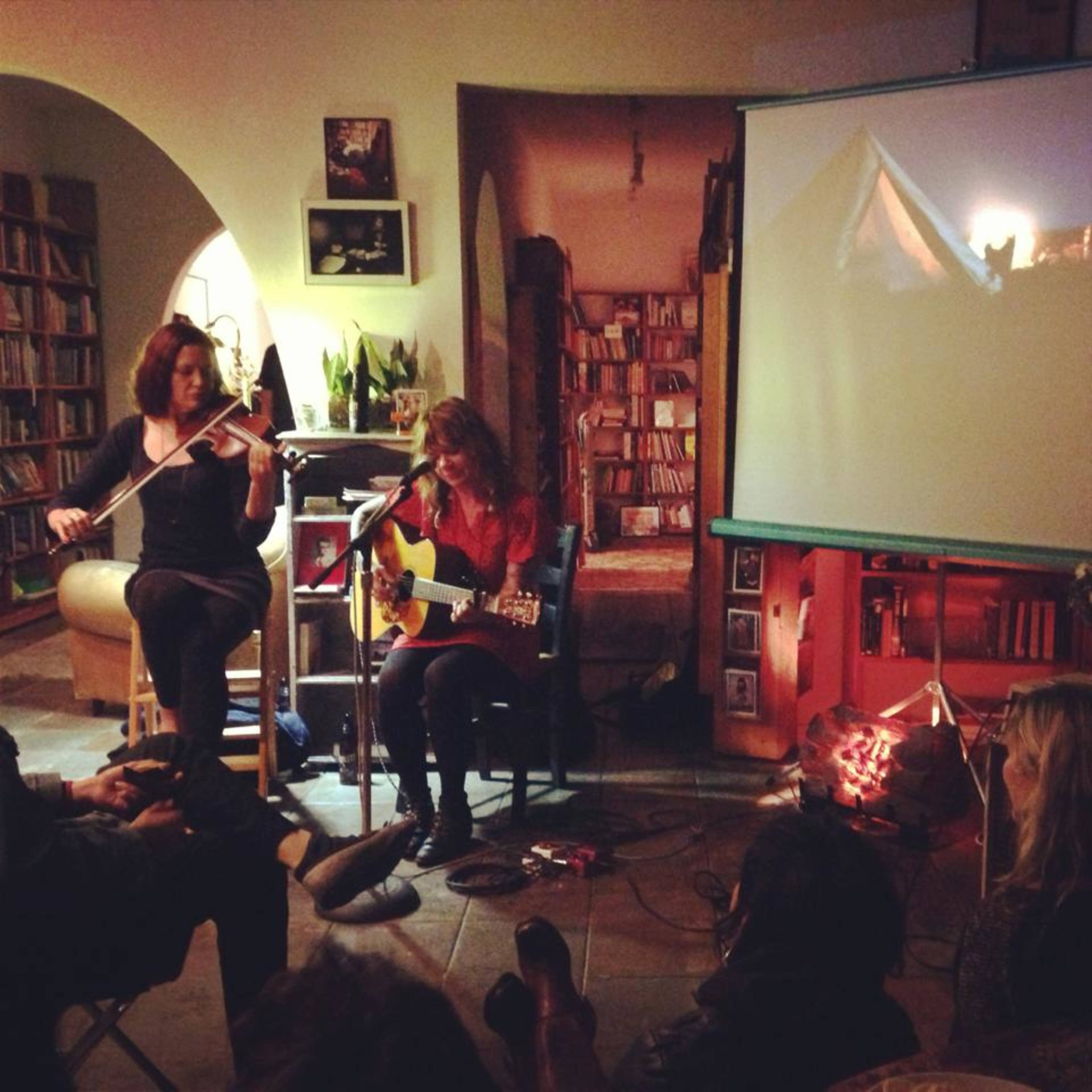 A woman plays guitar and sings on a small stage while another woman accompanies her on the violin, with a projector displaying an image on the wall behind them.