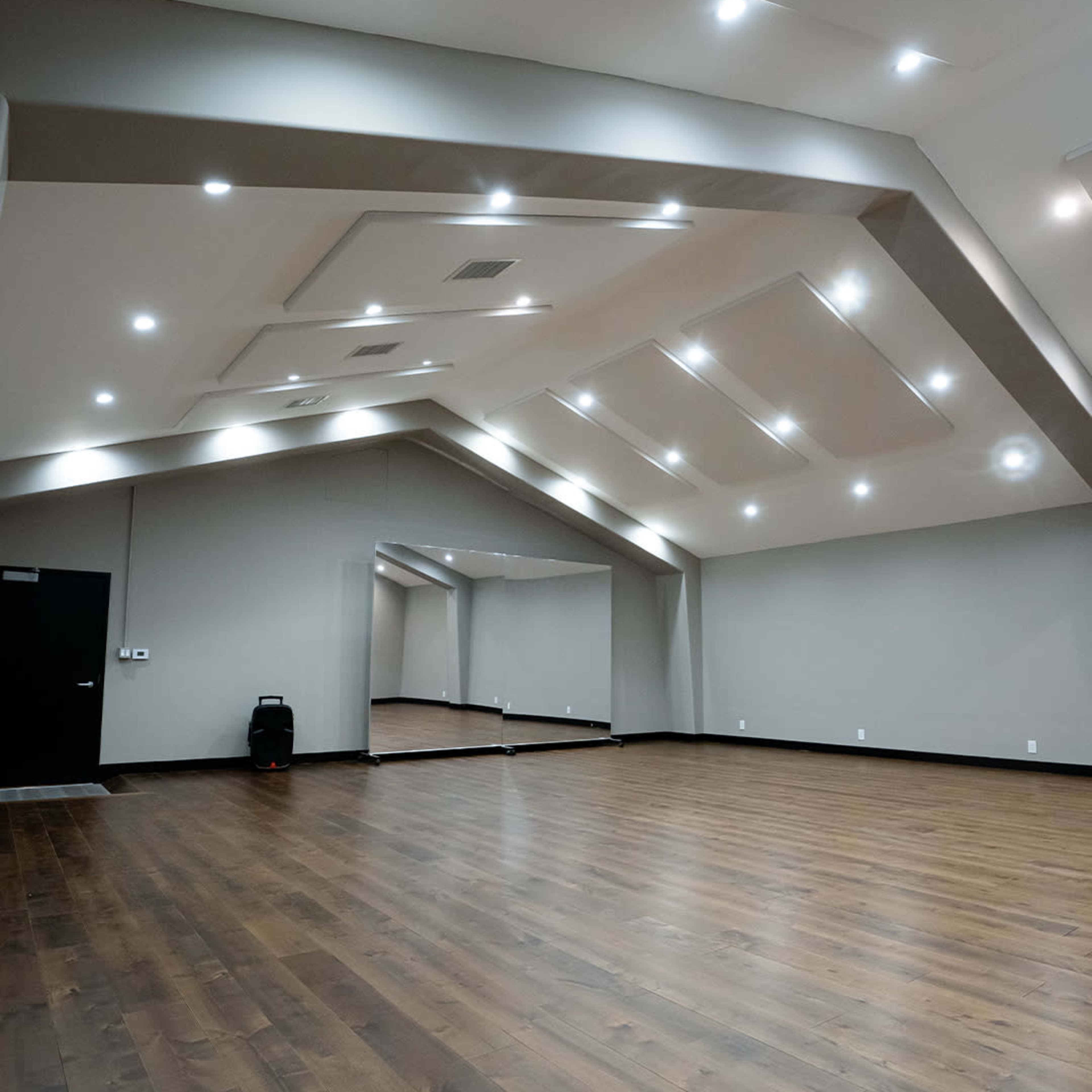 An empty dance studio with wooden flooring, high ceilings, and wall mirrors illuminated by recessed lighting.