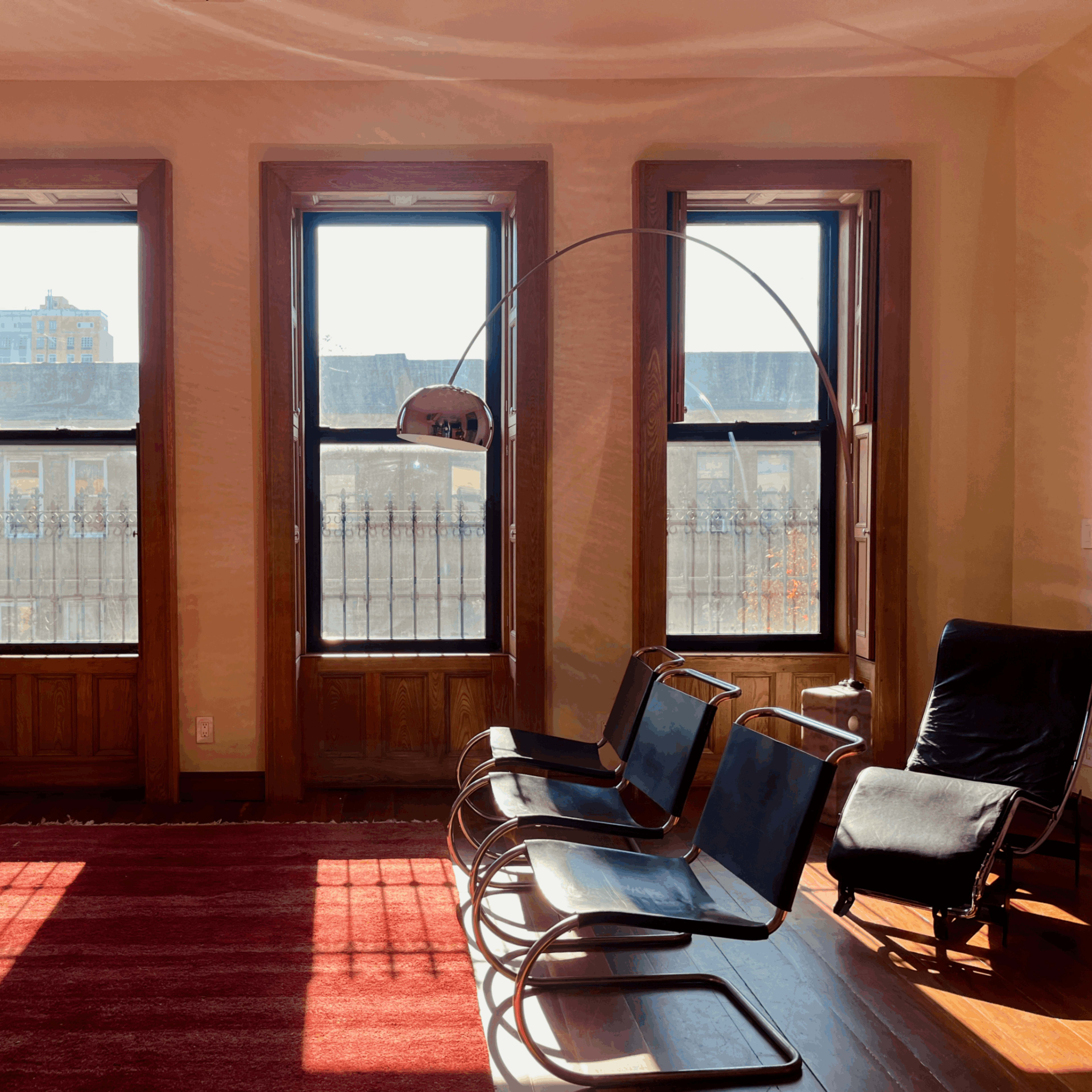 The scene features a sunlit room with three large windows, a dark sofa, two black chairs, and a red rug on the wooden floor.