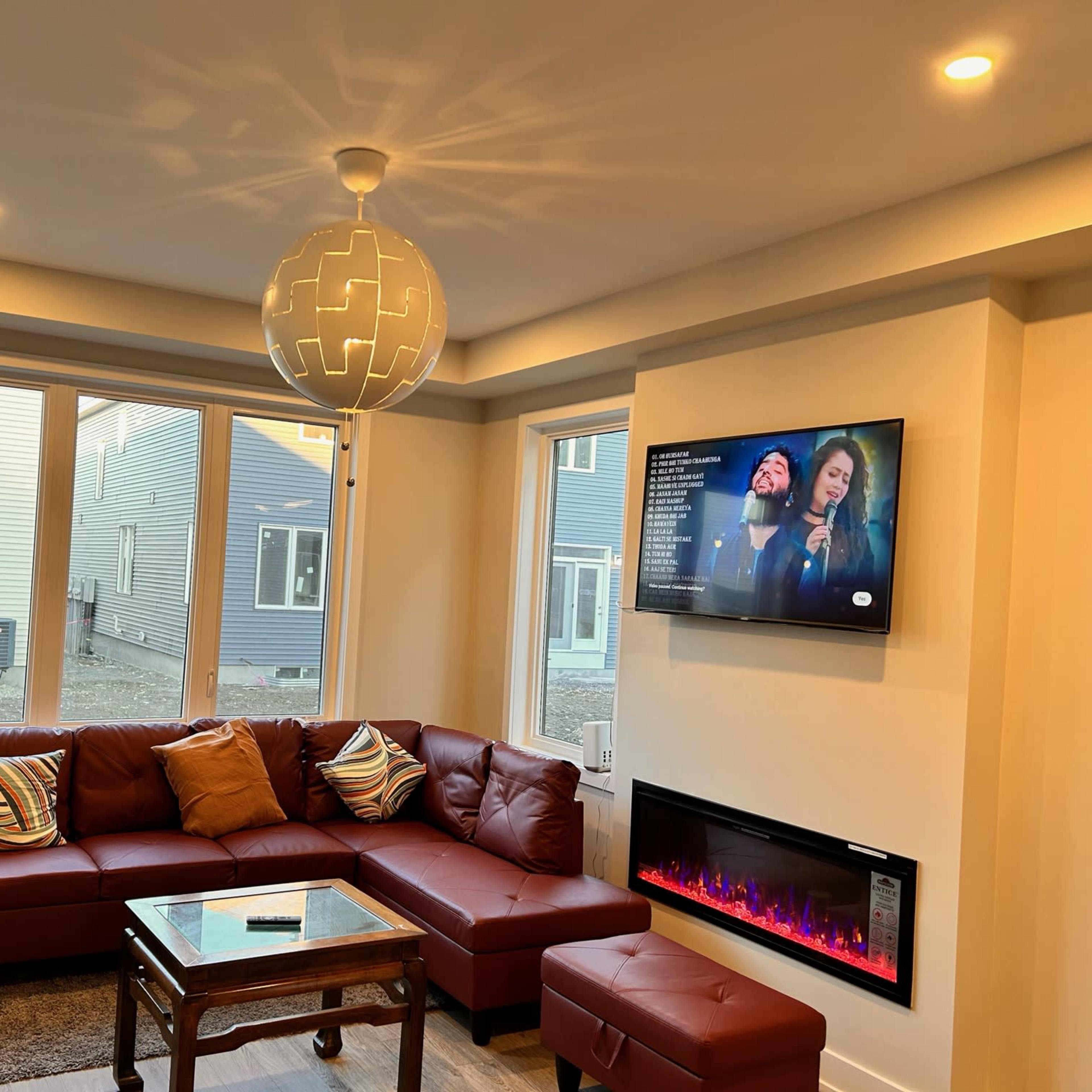 A modern living room featuring a brown sectional sofa, a wall-mounted television displaying a show, and a contemporary light fixture above a glass coffee table.