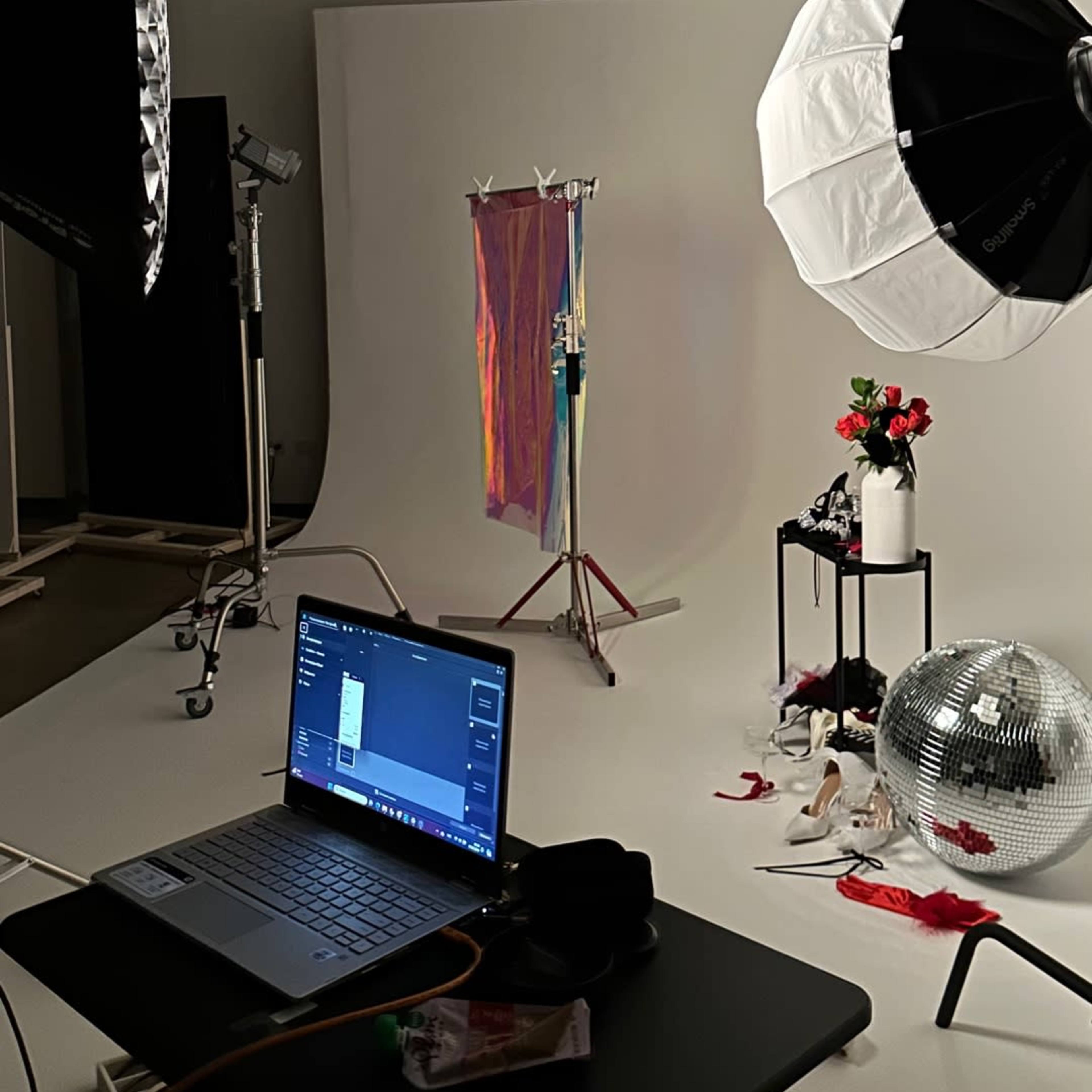 A photography studio set up with lighting equipment, a clothing rack draped with fabric, a laptop, and a disco ball on the floor.
