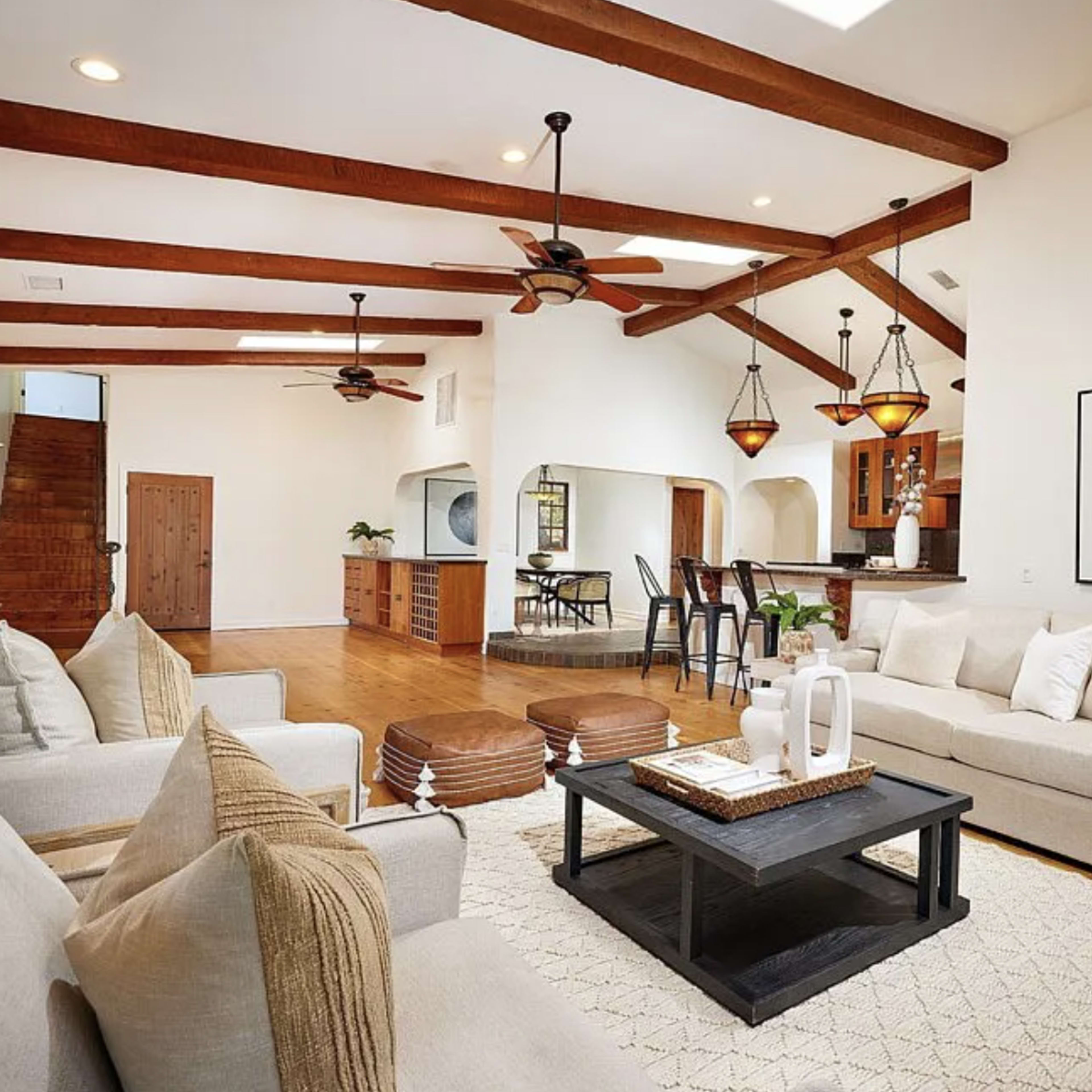 A spacious living room features light-colored sofas, a black coffee table, wooden beams on the ceiling, and a view into an adjacent kitchen area.