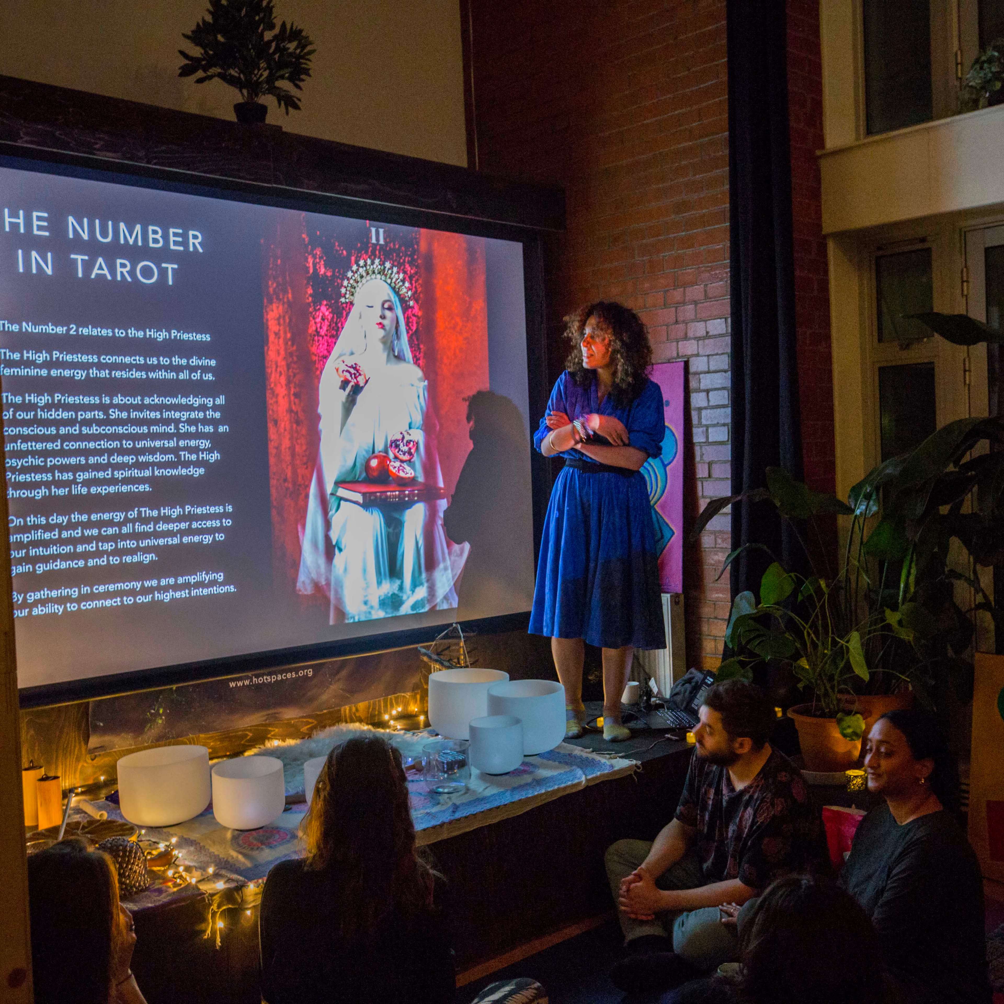 A woman in a blue dress stands in front of a large screen displaying information about the number 3 in Tarot, while an audience sits on the floor, surrounded by plants and decorations.