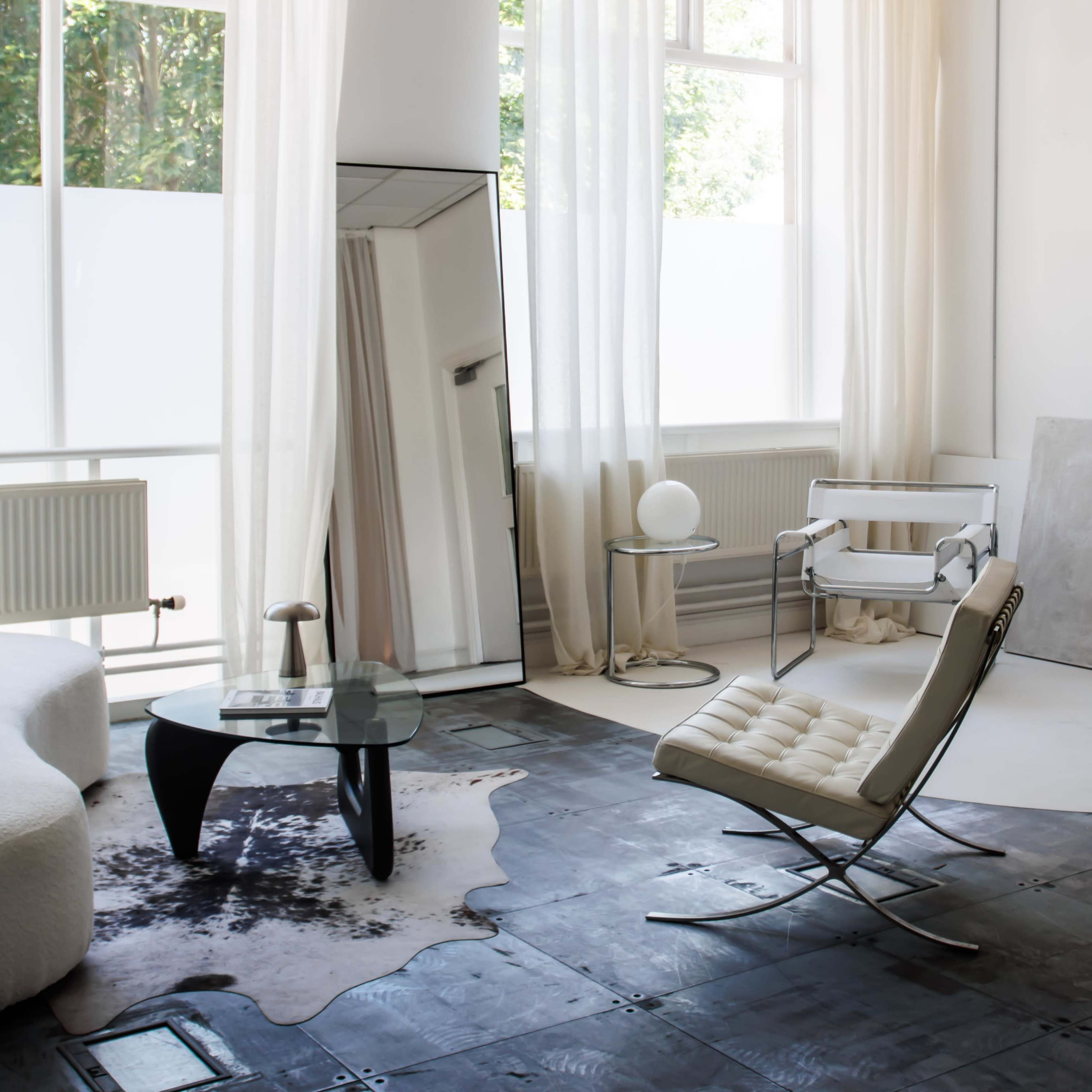 A bright, minimalistic living space features a white upholstered sofa, a glass coffee table, and a contemporary lounge chair, with large windows allowing ample natural light.