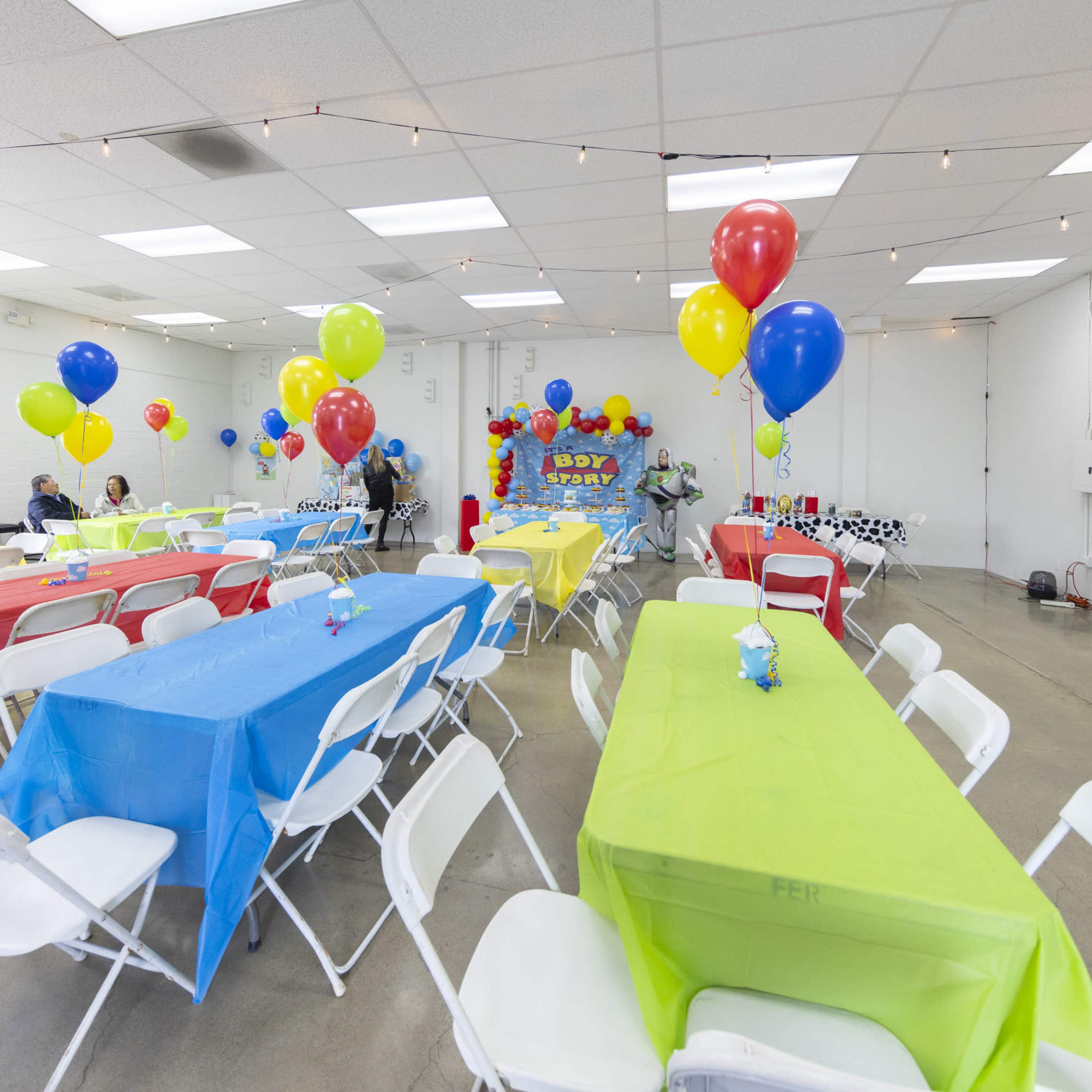 A spacious party room is decorated with colorful tablecloths, balloons, and a festive sign, featuring several seating arrangements for guests.