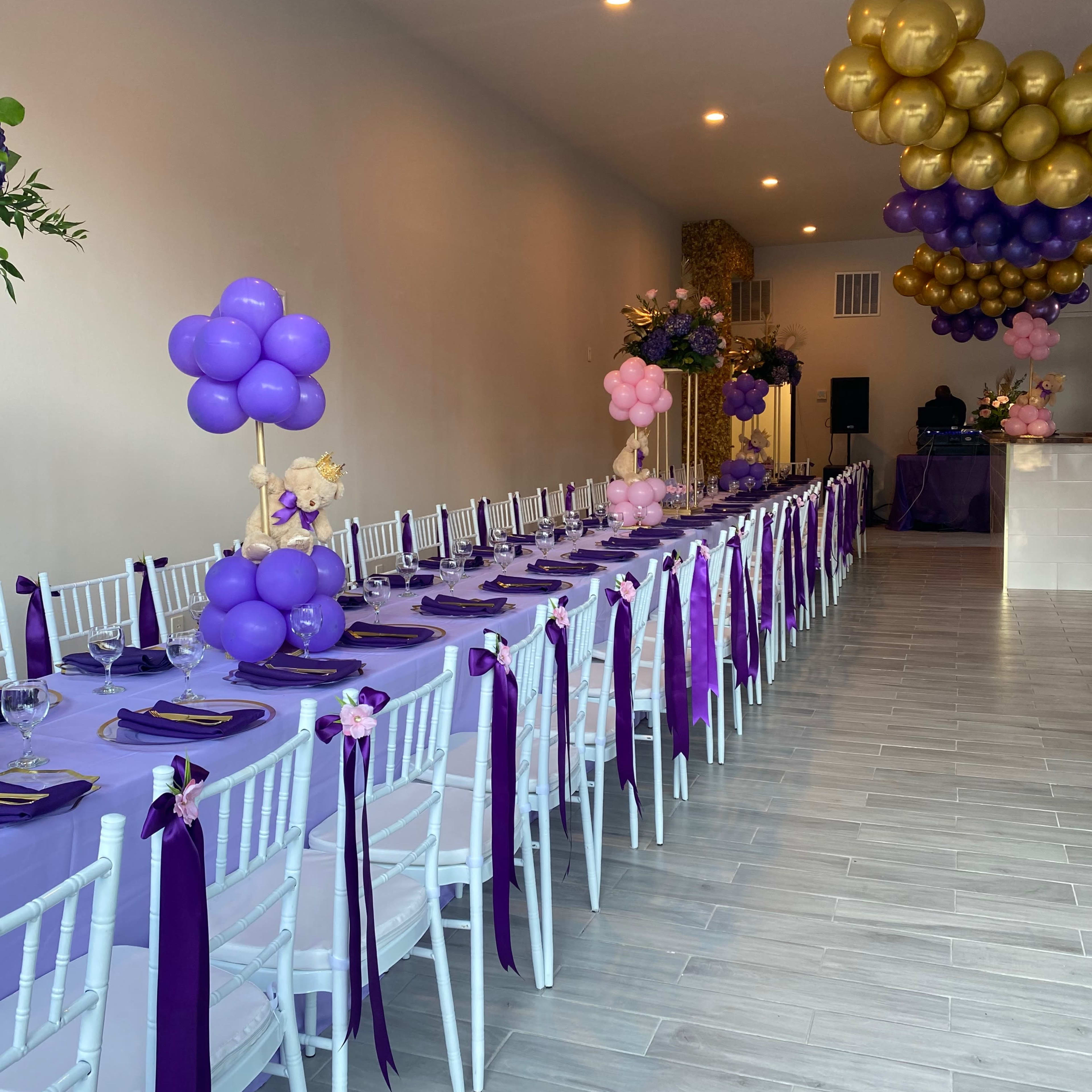 A long table with purple linens and white chairs, decorated with balloon centerpieces and set for a celebration.