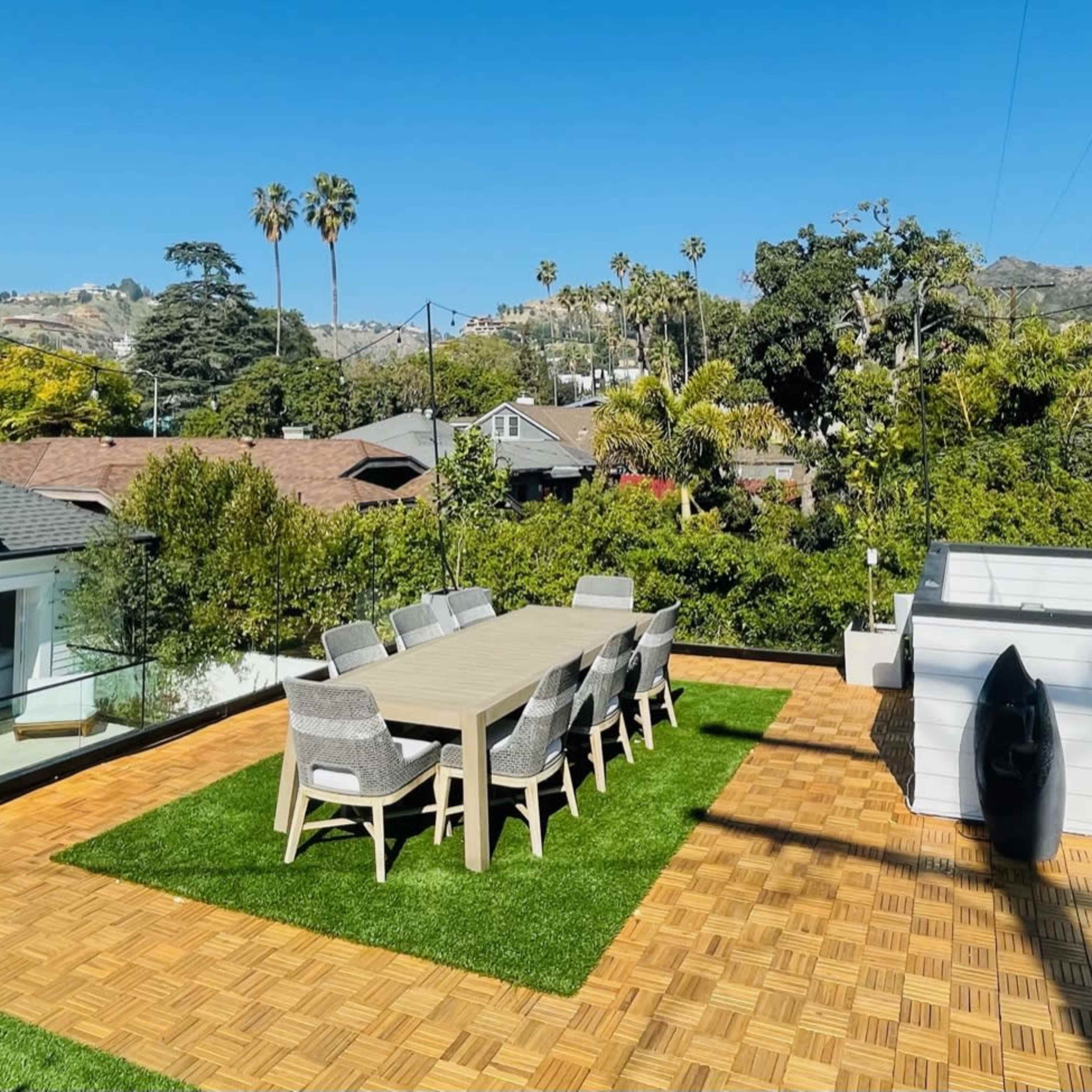 A rooftop terrace featuring a large dining table surrounded by chairs, artificial grass, and views of palm trees and distant hills.