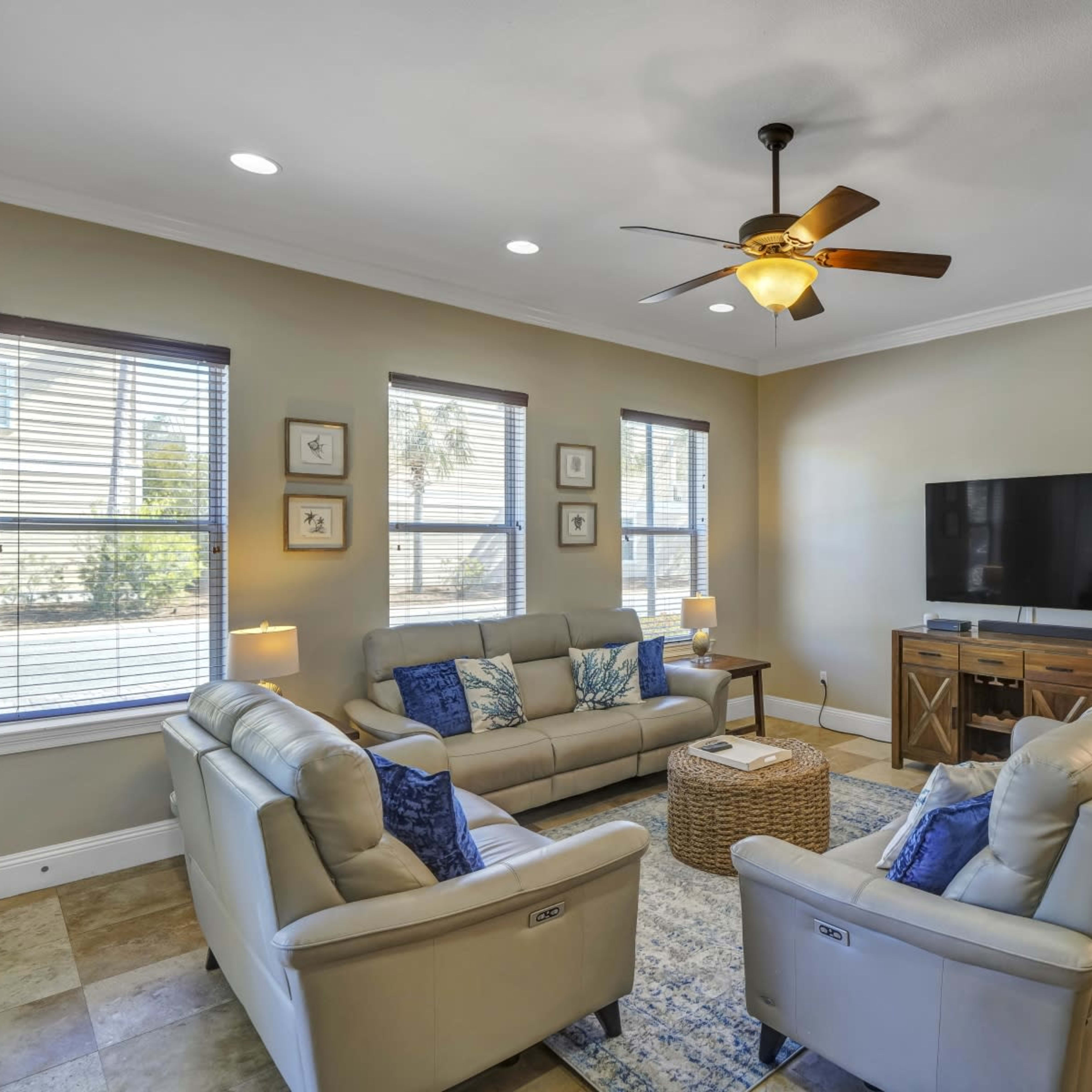 The living room features two couches facing a television, with a coffee table and a woven rug in the center, illuminated by natural light from the windows.