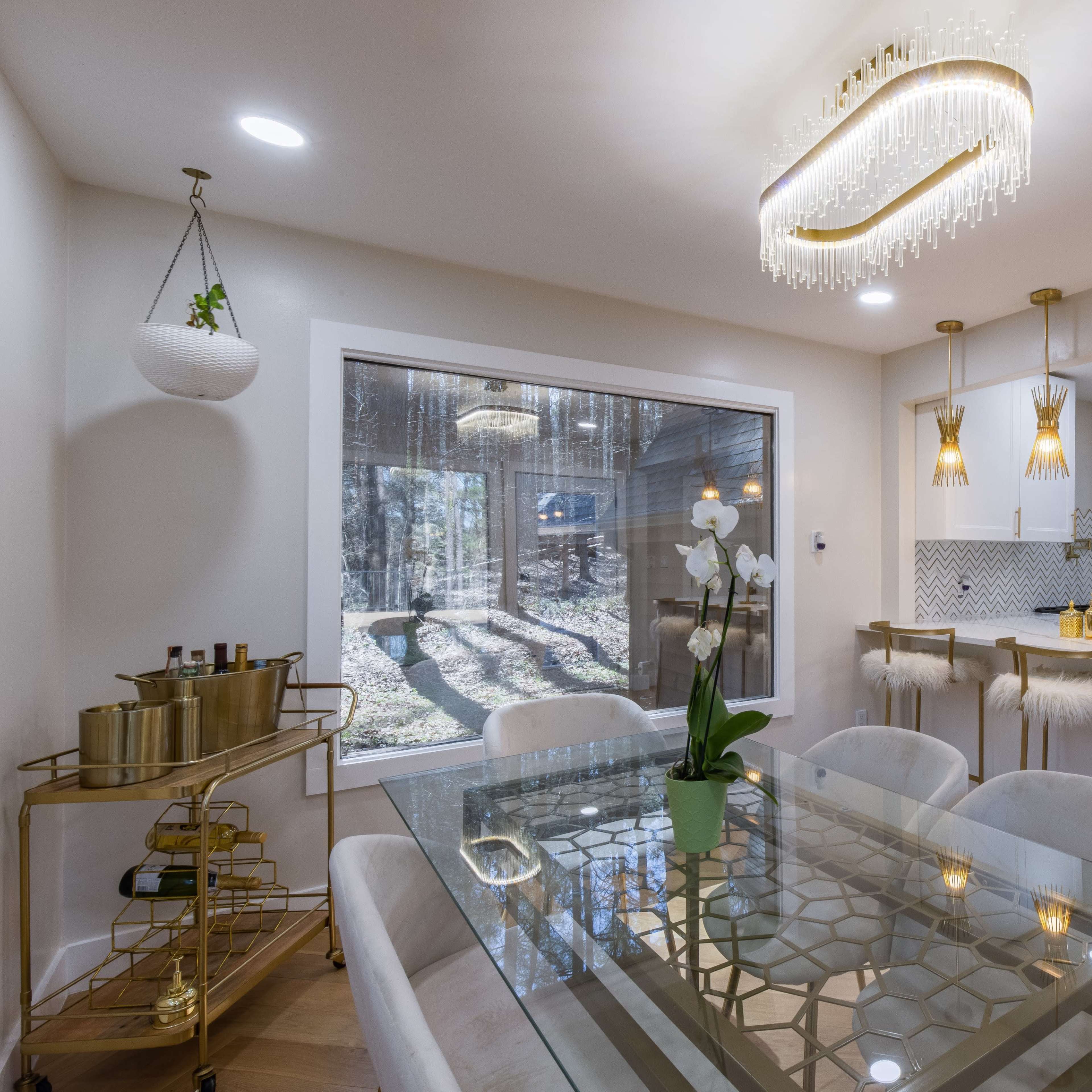 A modern dining area features a glass table with six upholstered chairs, a gold bar cart, and a bright chandelier above, with a view of trees through a large window.