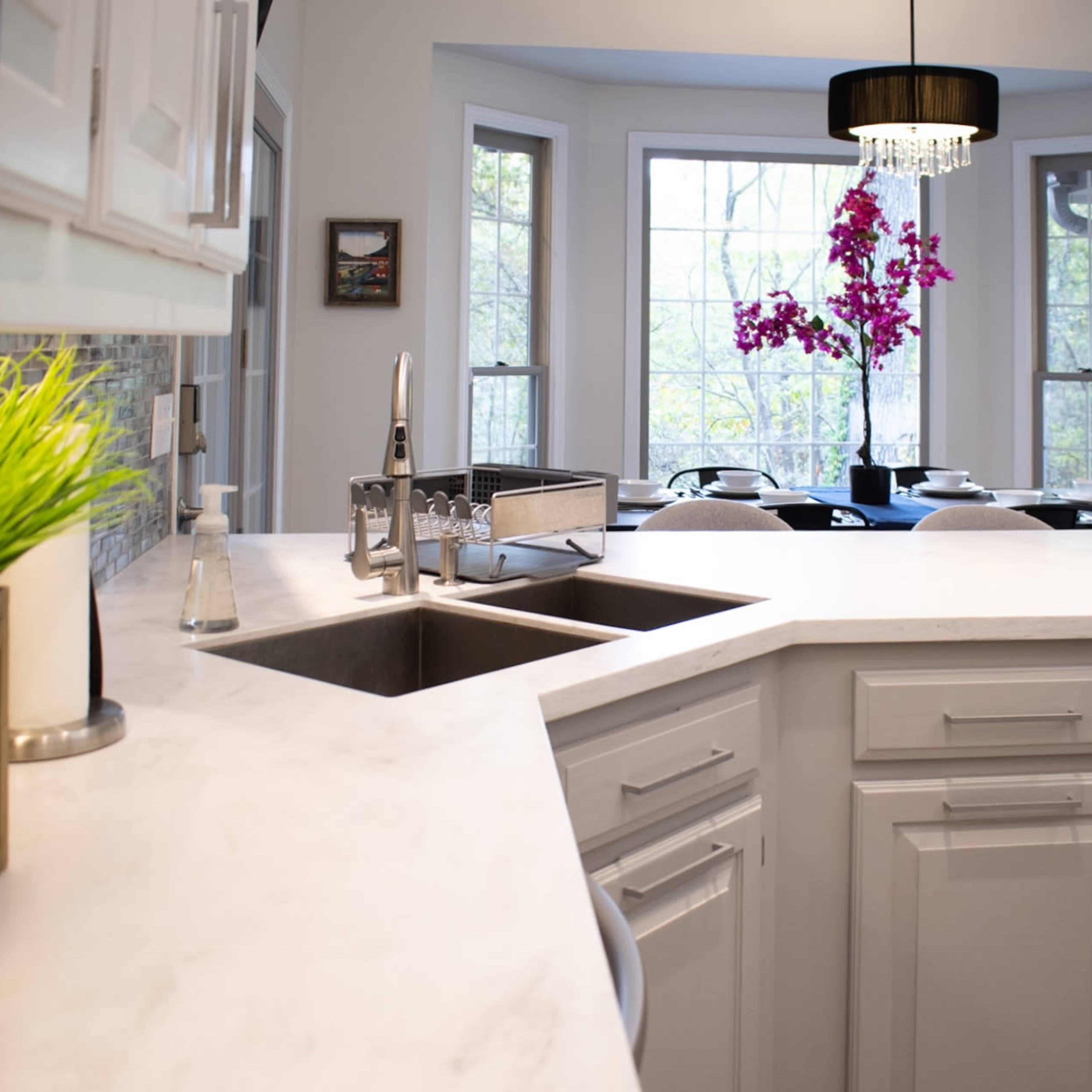 A modern kitchen features white cabinets, a marble countertop with a double sink, and a bay window with a view of a dining area adorned with colorful flowers.