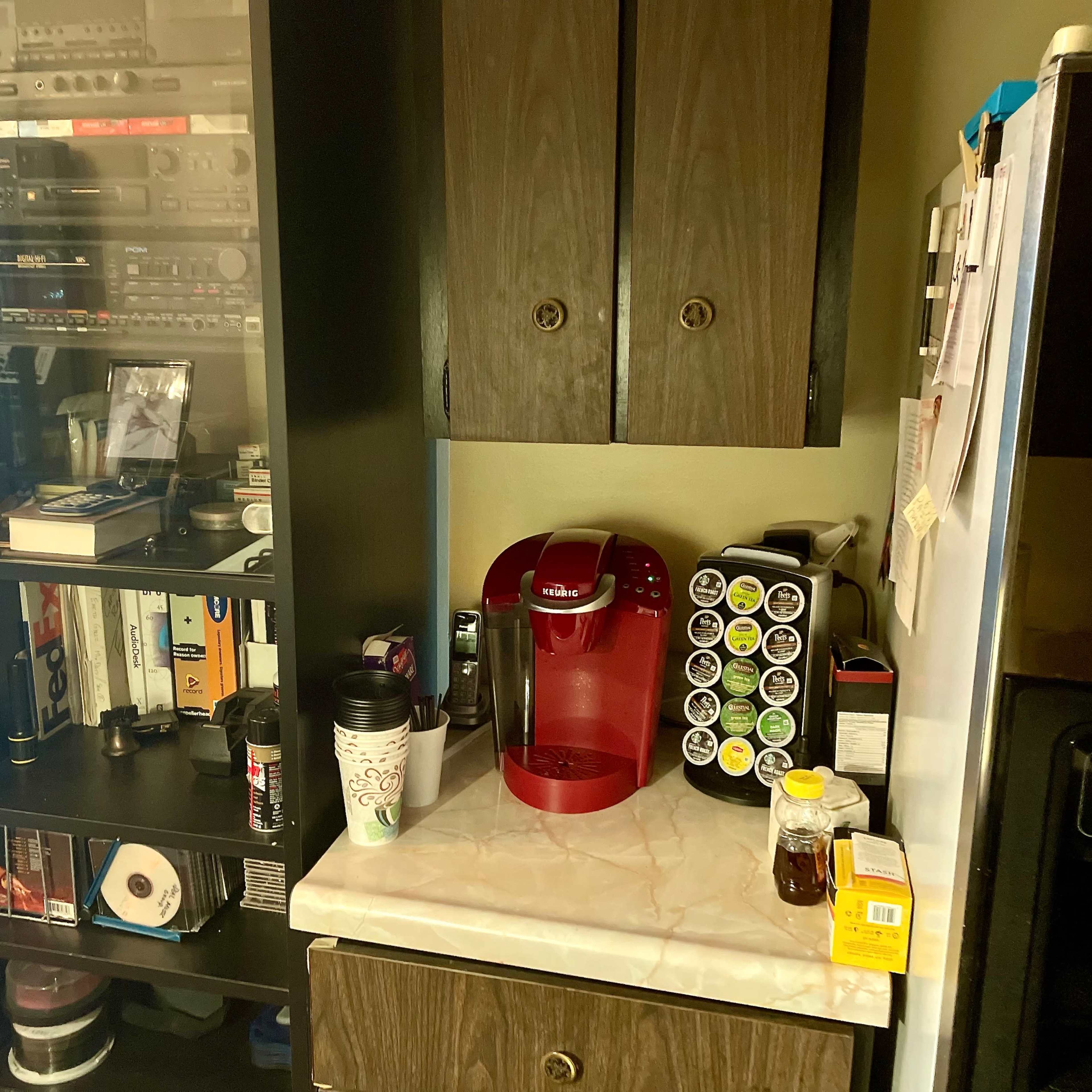 A kitchen corner featuring a coffee maker, a spice rack, and a shelving unit with various media equipment and books.