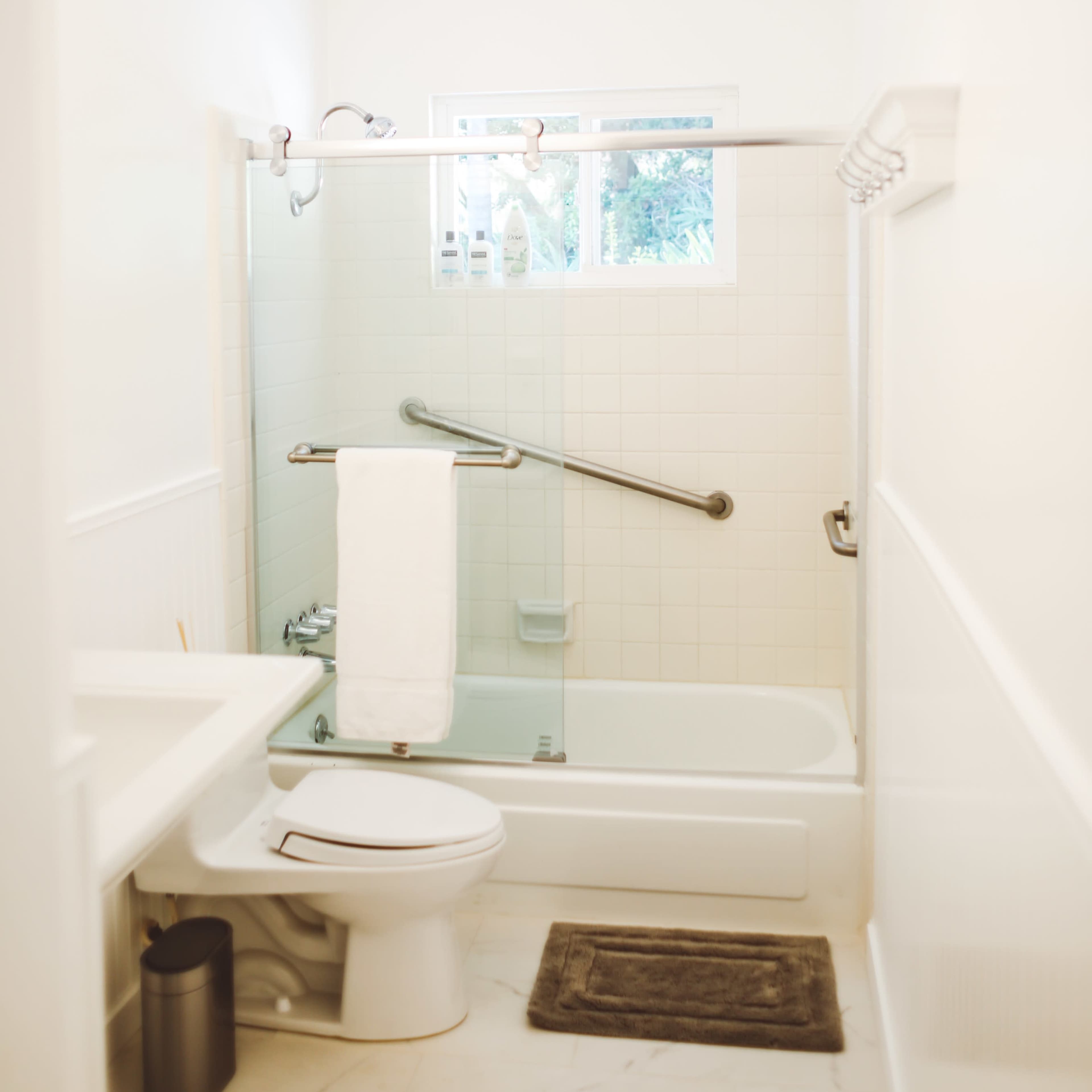 A clean, white-tiled bathroom with a bathtub, toilet, and a window providing natural light.