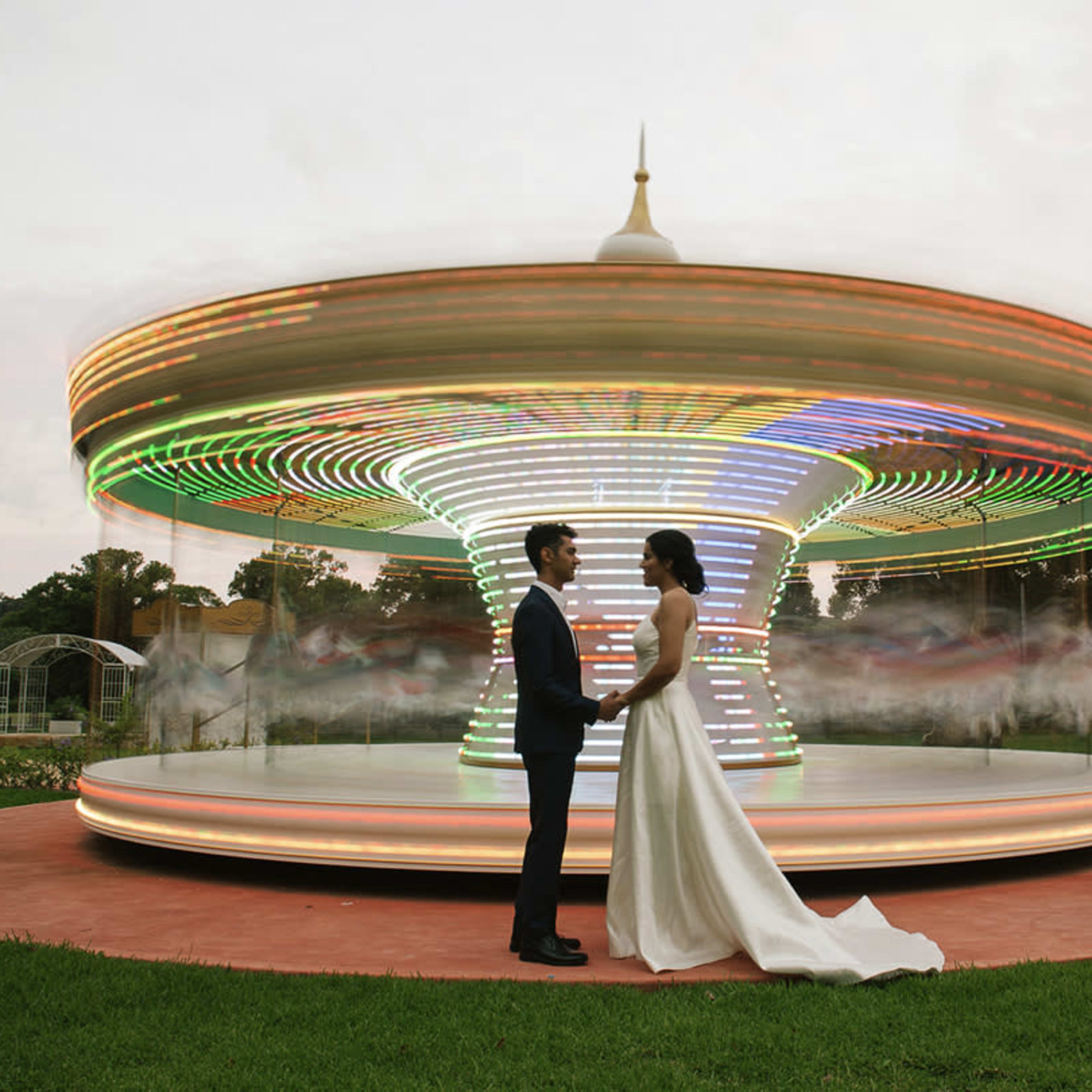A couple stands facing each other in front of a brightly lit carousel in a green park.