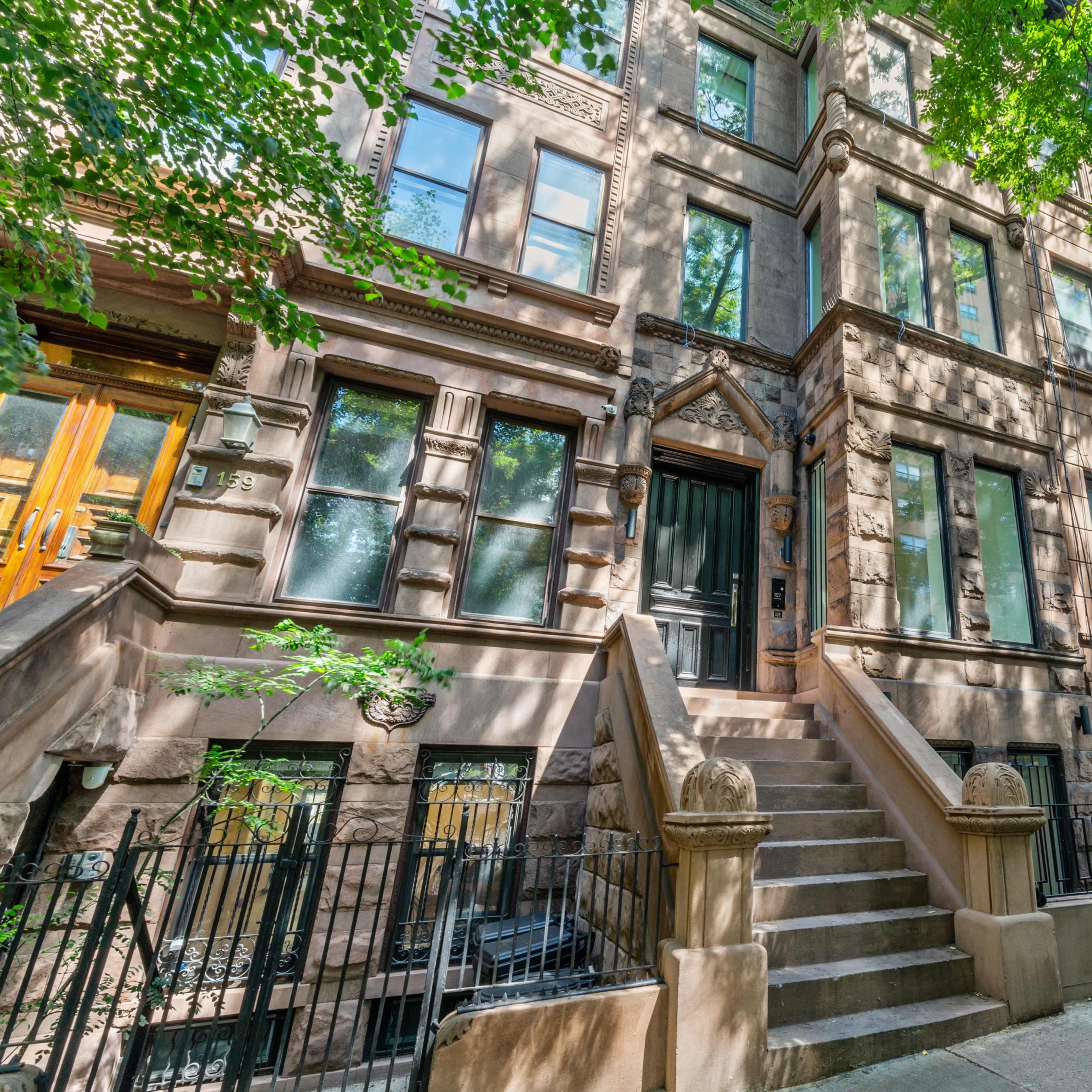 A row of brownstone buildings with intricate architectural details is lined along a sidewalk shaded by large trees.