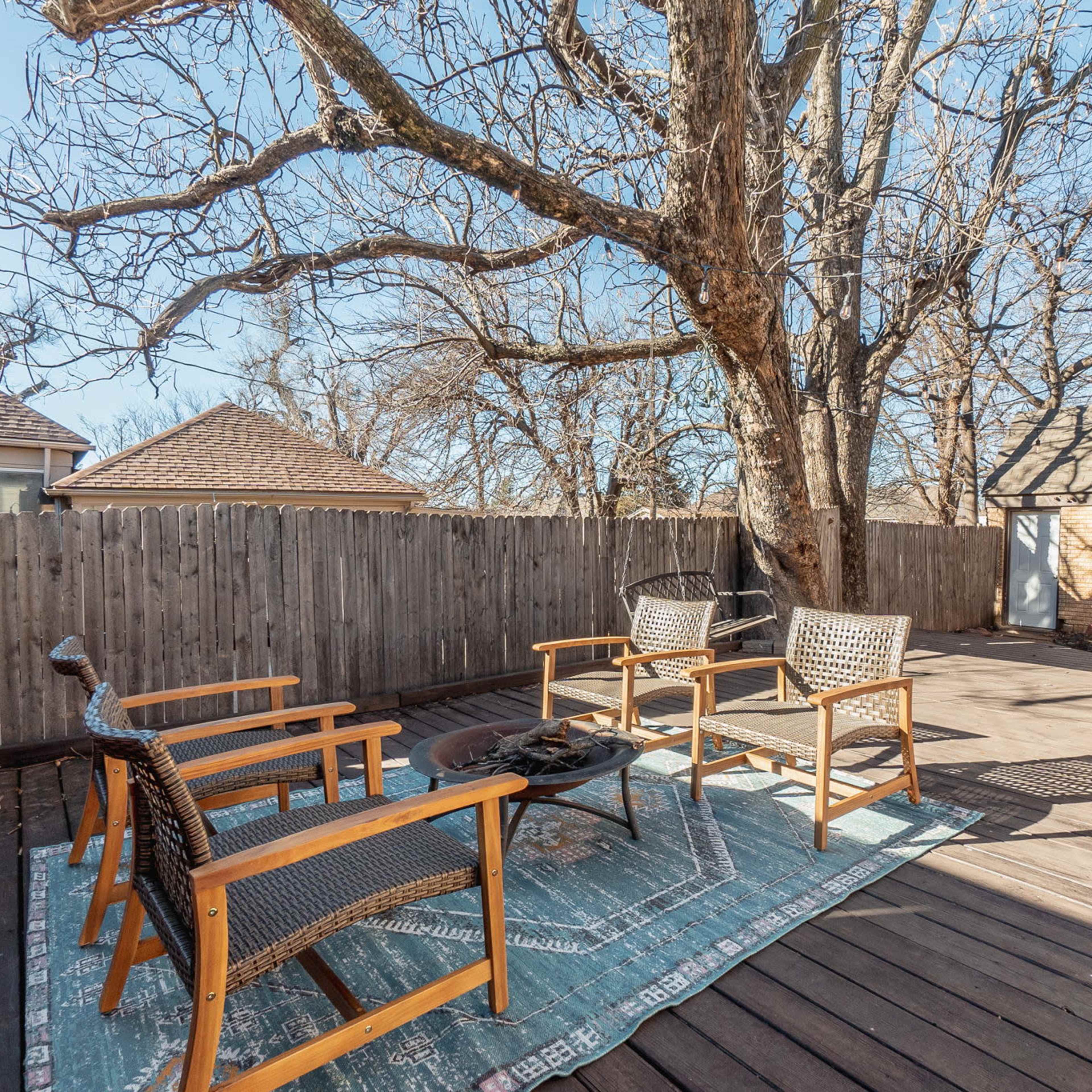 A wooden deck features four chairs arranged around a small table, with a large tree and a fence in the background.