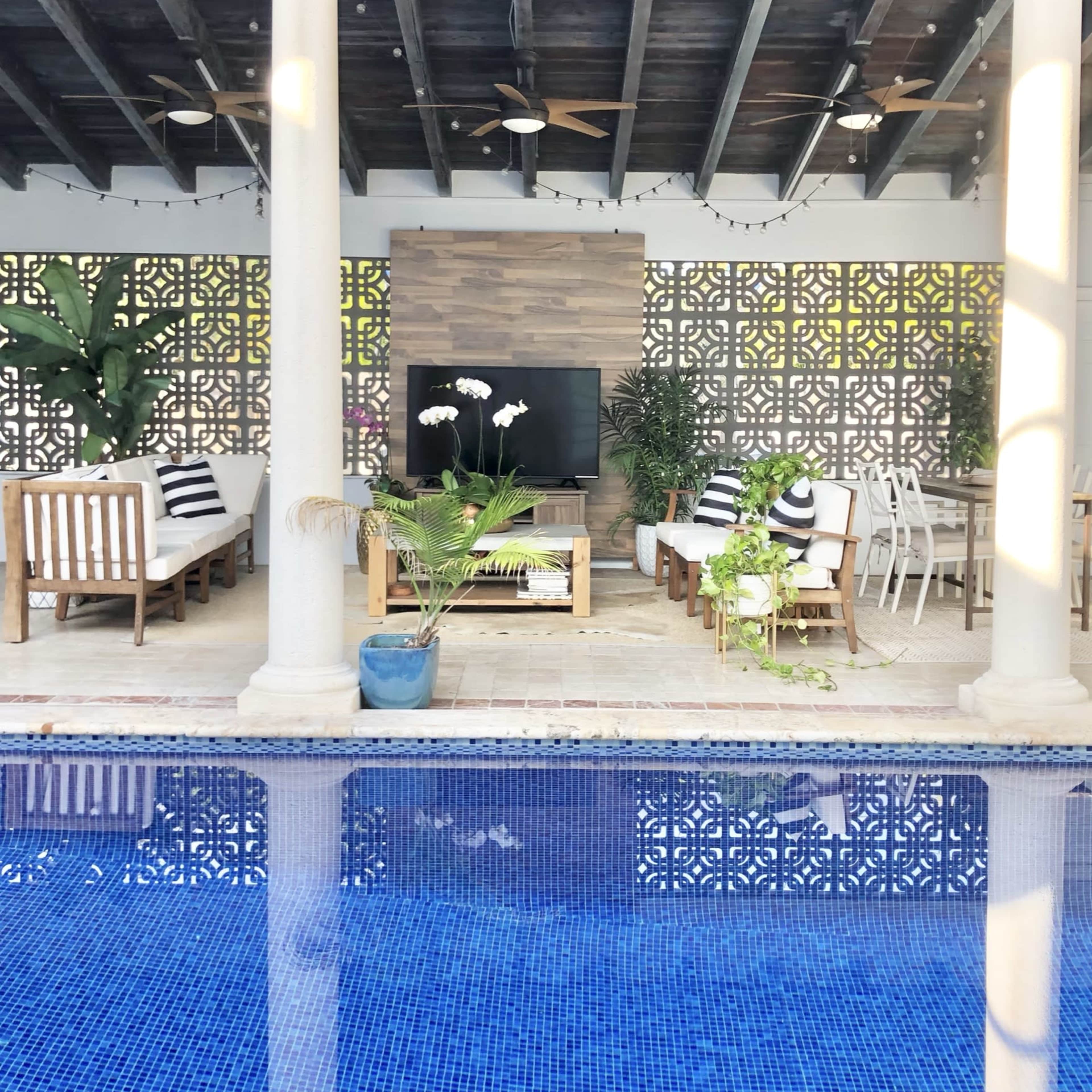 A patio area with seating, a television, and dining space overlooks a blue swimming pool.