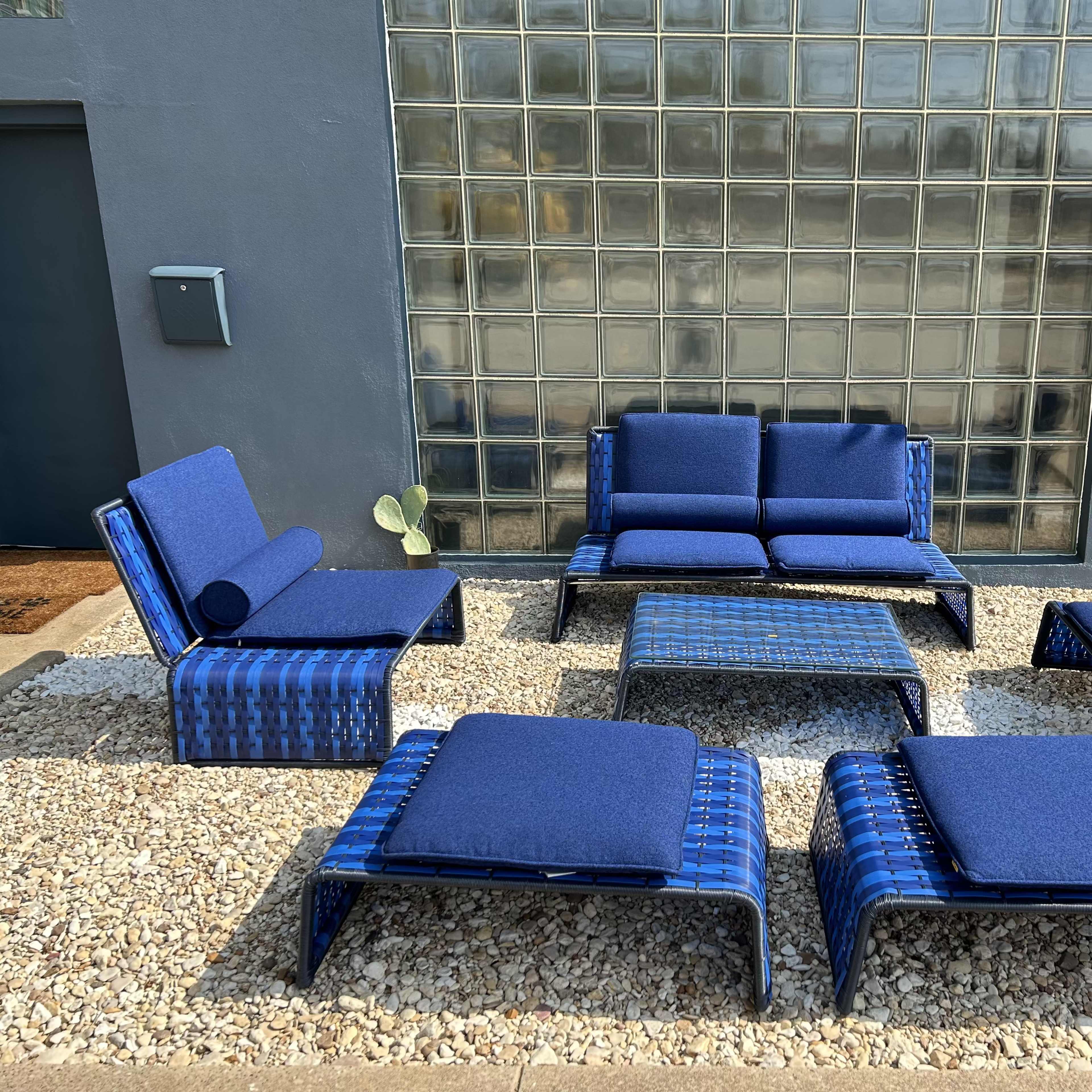 A set of six outdoor lounge chairs and a table, all in navy blue, are arranged on a pebble-strewn patio with a glass block wall in the background.