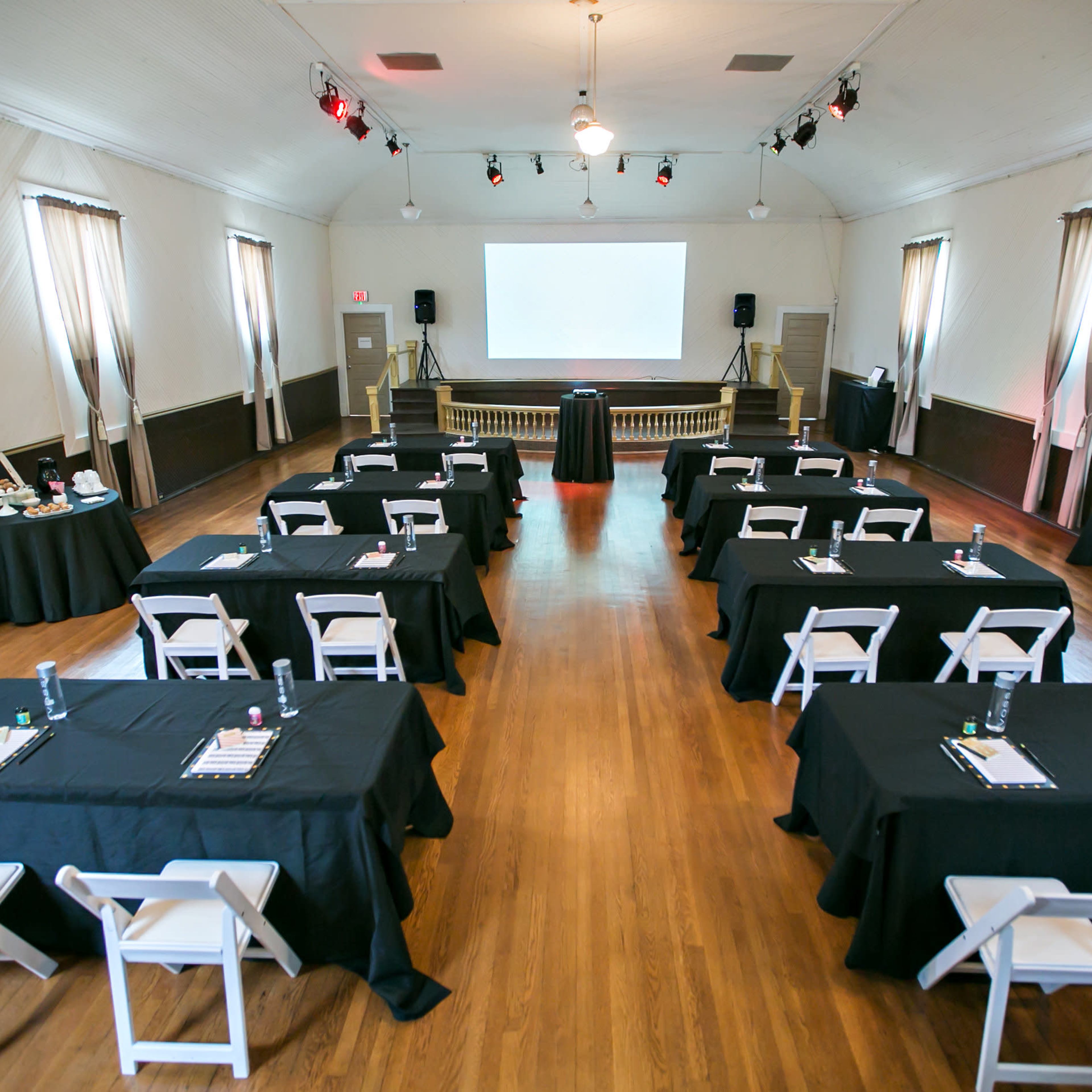 A spacious conference room set up with several tables arranged in a U-shape, each covered with black cloth and accompanied by chairs, facing a central presentation area with a projector screen.