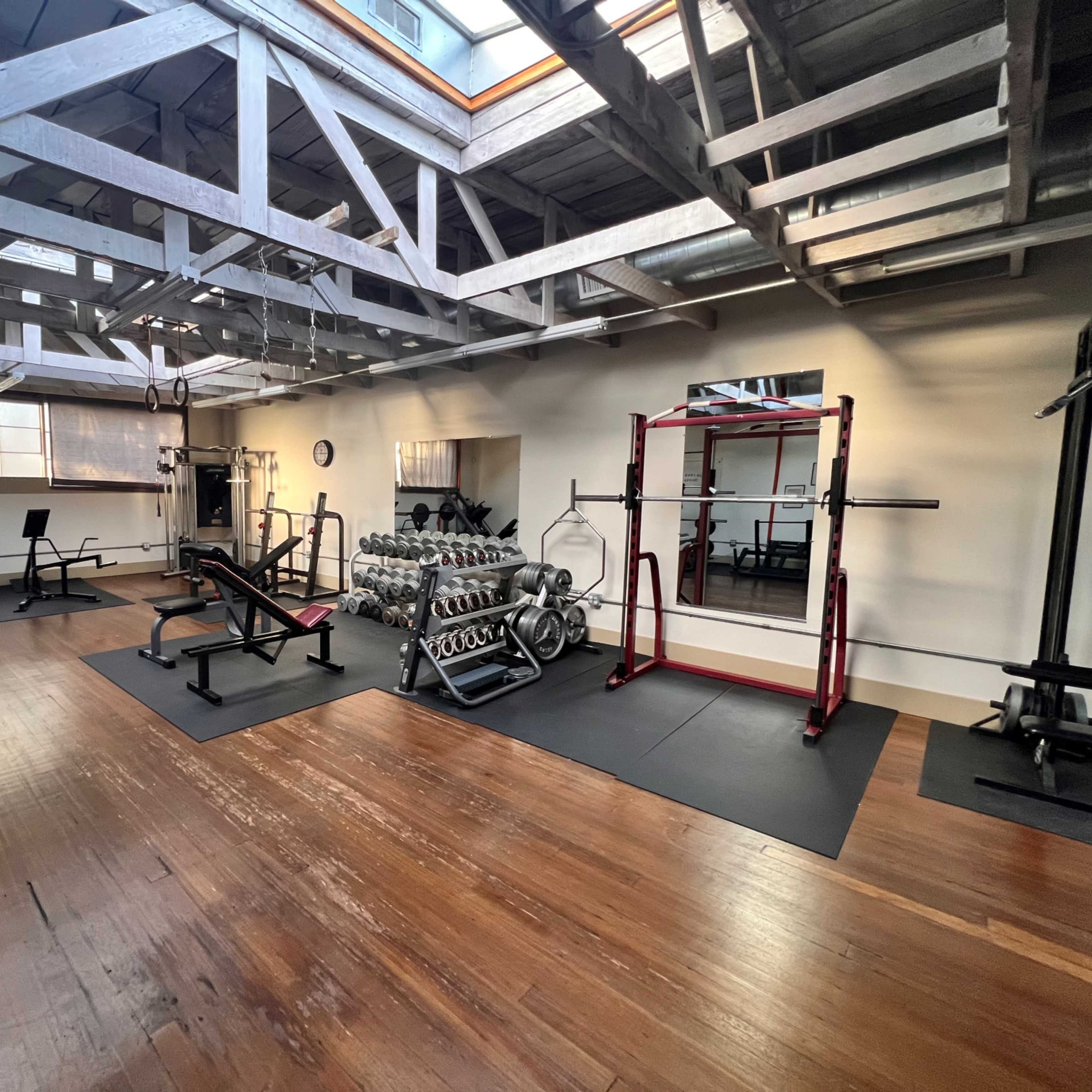 A gym with various weightlifting equipment, benches, and a rack of dumbbells on a wooden floor beneath a ceiling supported by wooden beams.