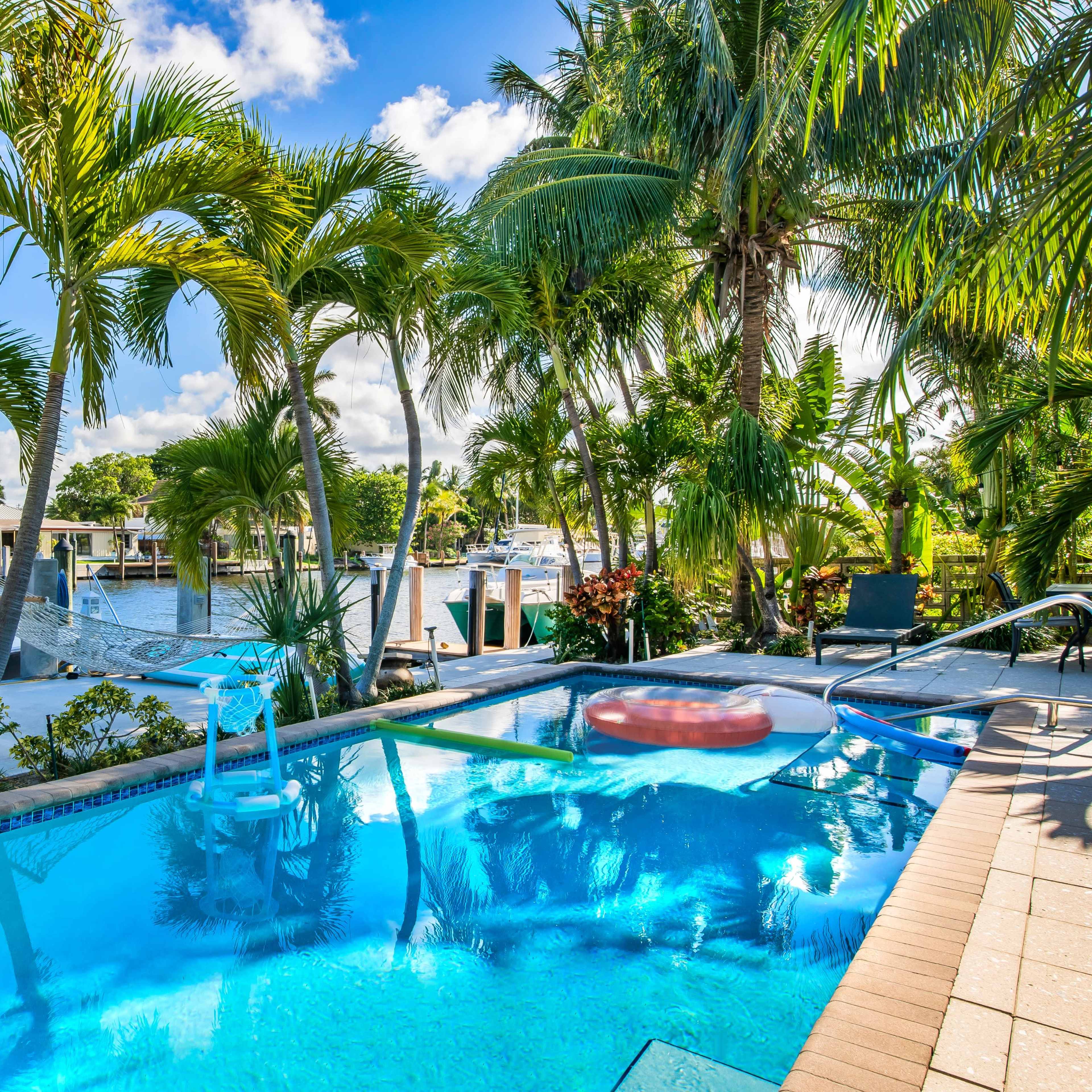 A swimming pool surrounded by palm trees and a waterfront view with boats docked in the background.