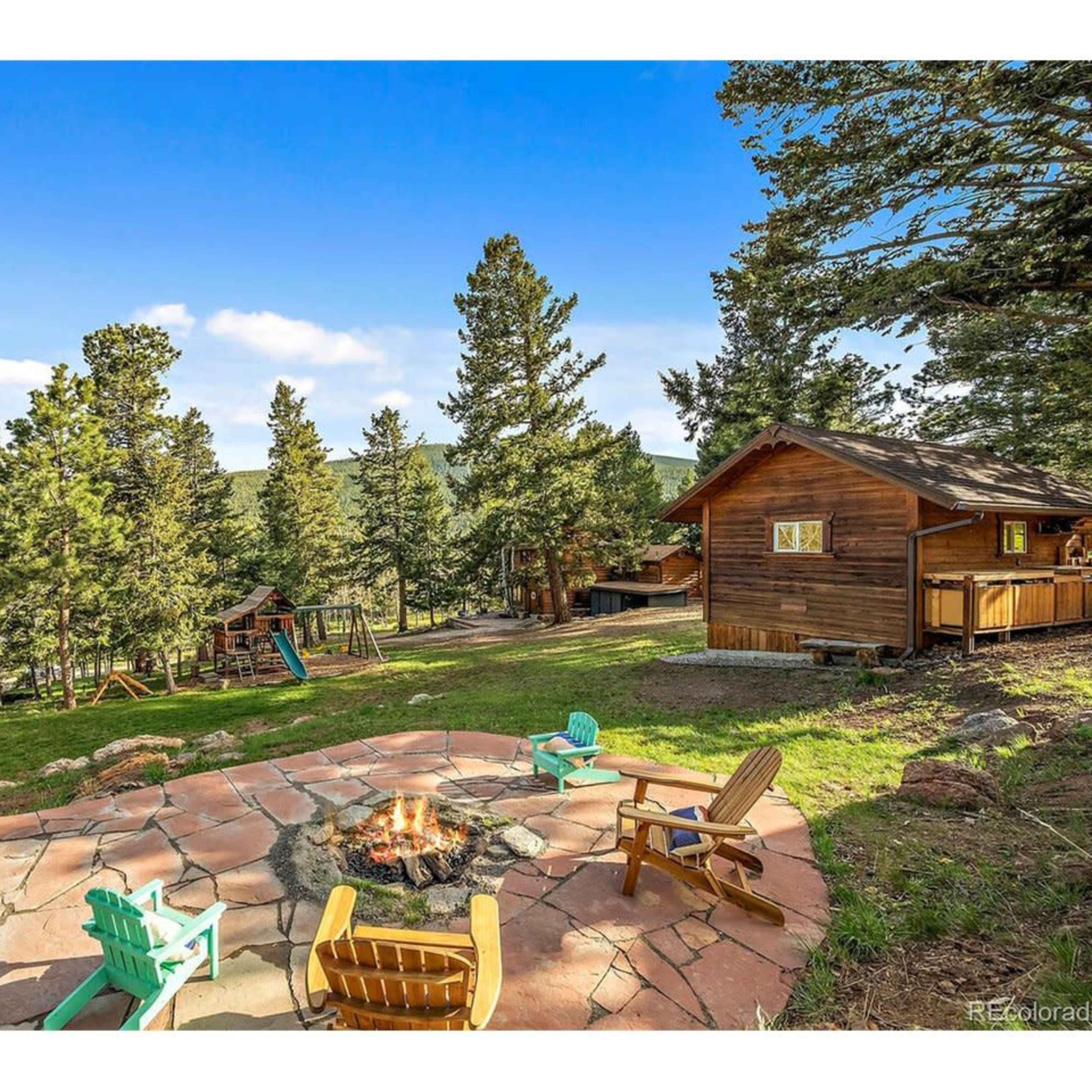 A stone patio with Adirondack chairs surrounds a fire pit, set against a backdrop of tall trees and a cabin in a mountainous area.