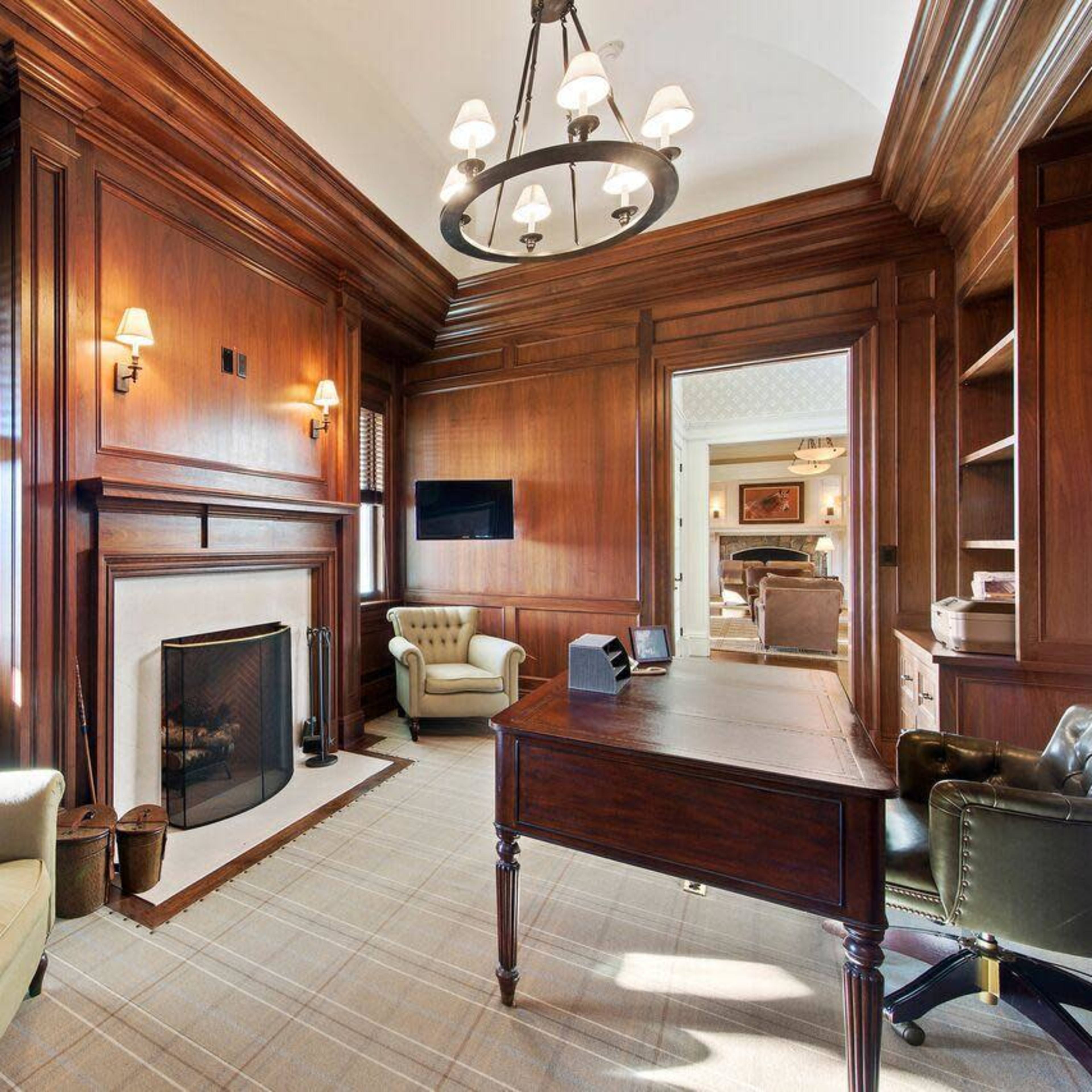 A wooden-paneled office featuring a fireplace, a desk, two armchairs, and a large window with scenic views.