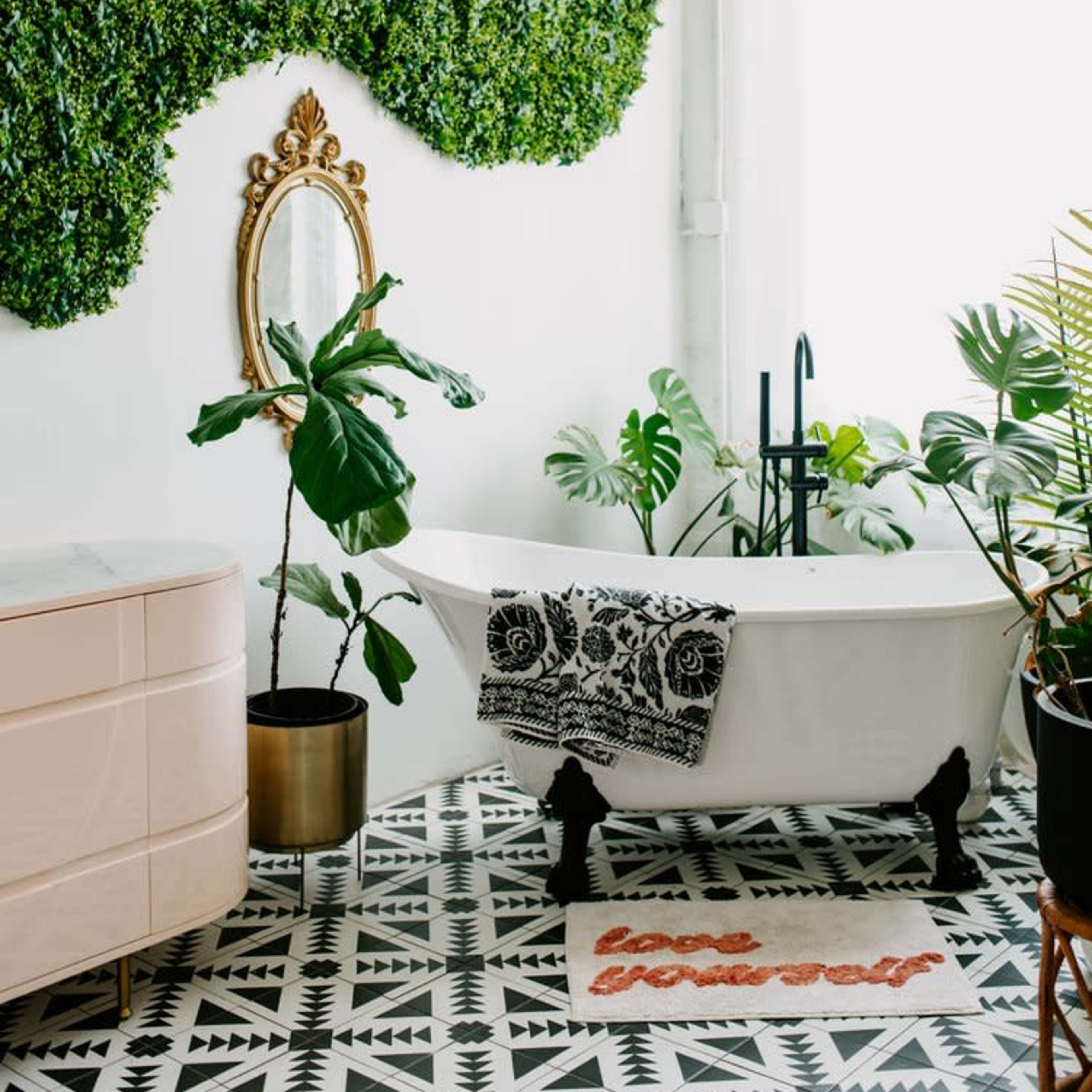 A bathroom featuring a freestanding white bathtub, a large green wall of artificial plants, and a patterned tiled floor with a decorative bath mat.