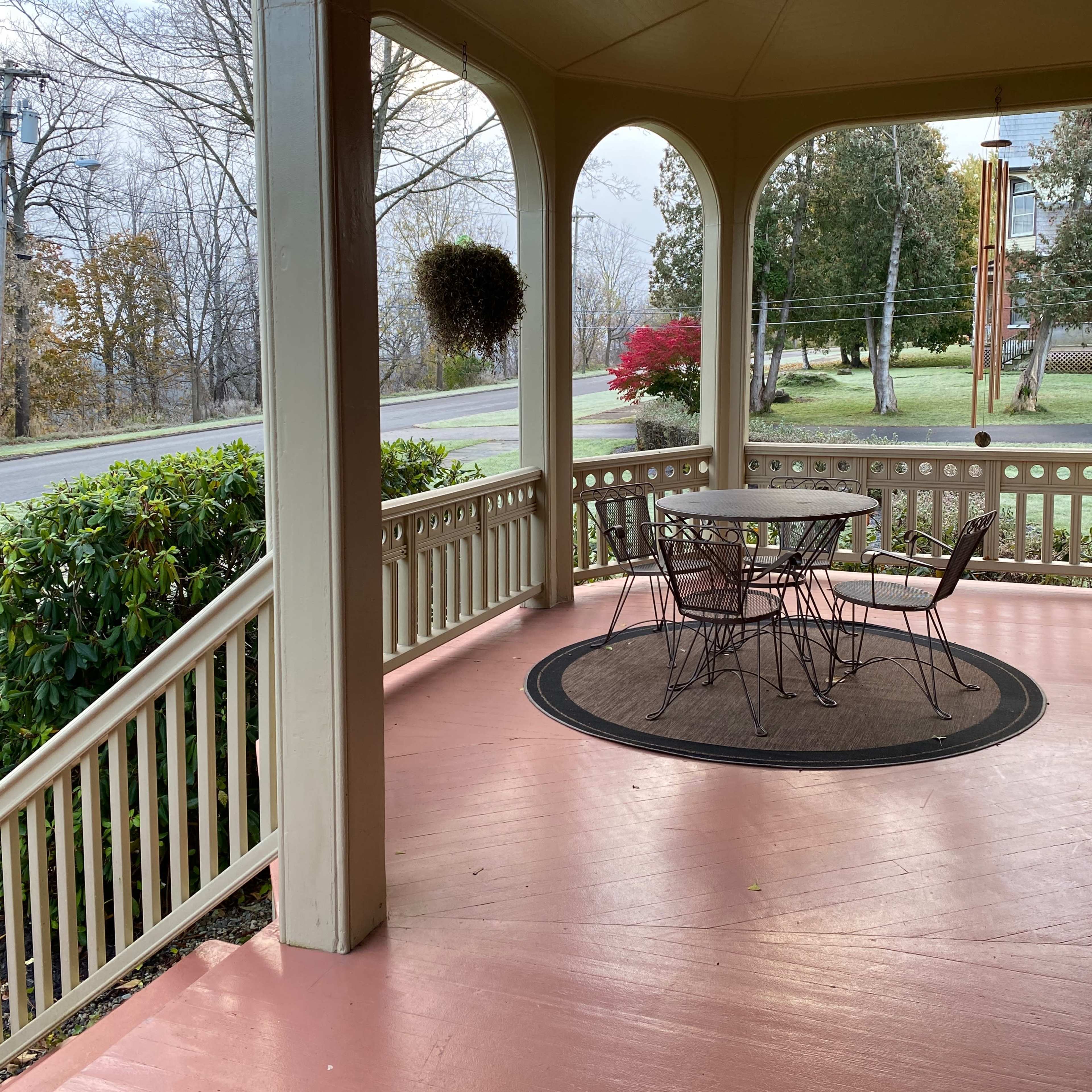 A porch with a round table and chairs, surrounded by a railing and potted plants, overlooking a quiet street.