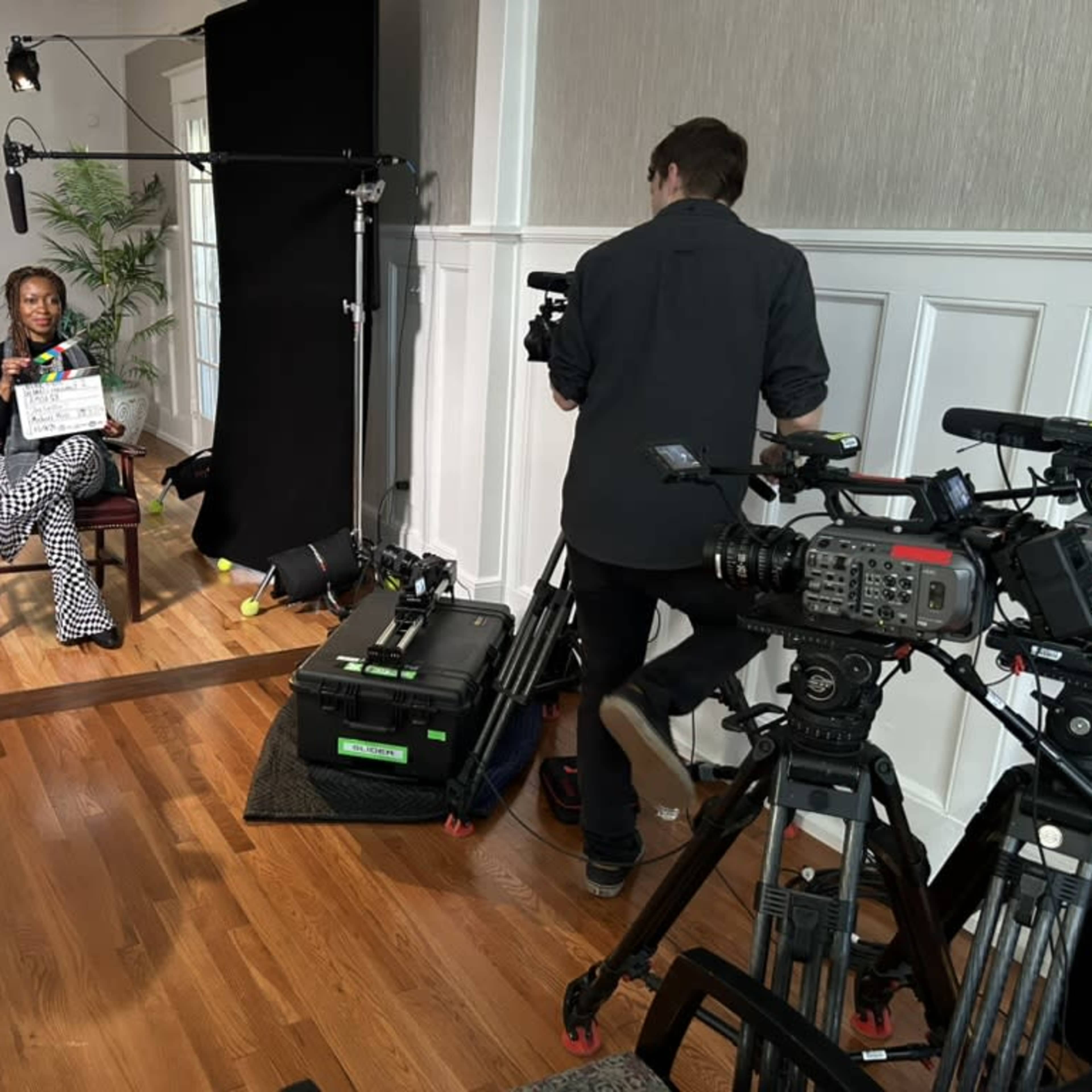 A woman sits in a chair holding a clapperboard while a man operates a camera nearby in a film set environment.