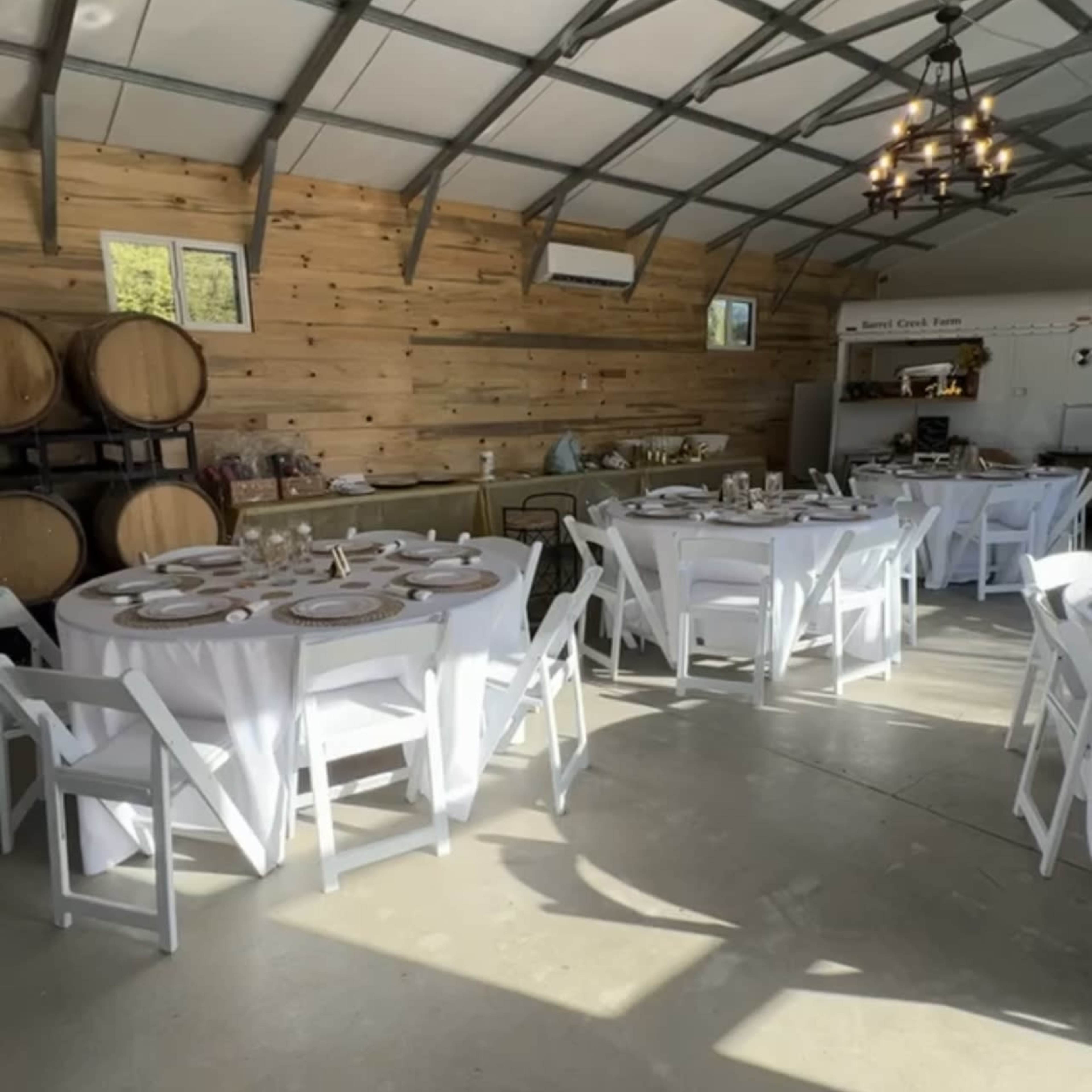 A rustic dining space with multiple round tables set with white tablecloths, surrounded by wooden walls and wine barrels.
