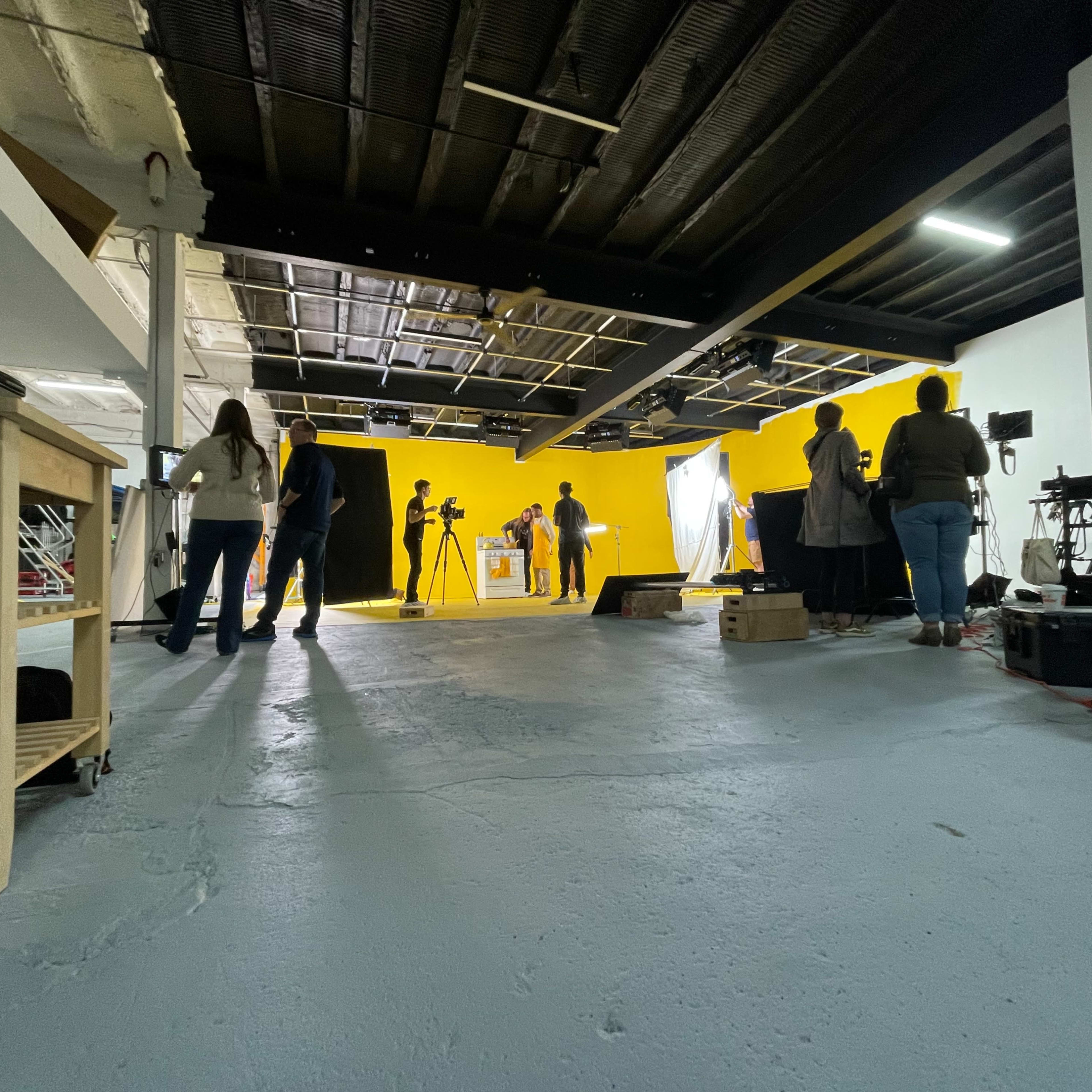 A photography studio features a bright yellow wall, with several people setting up equipment and preparing for a shoot.