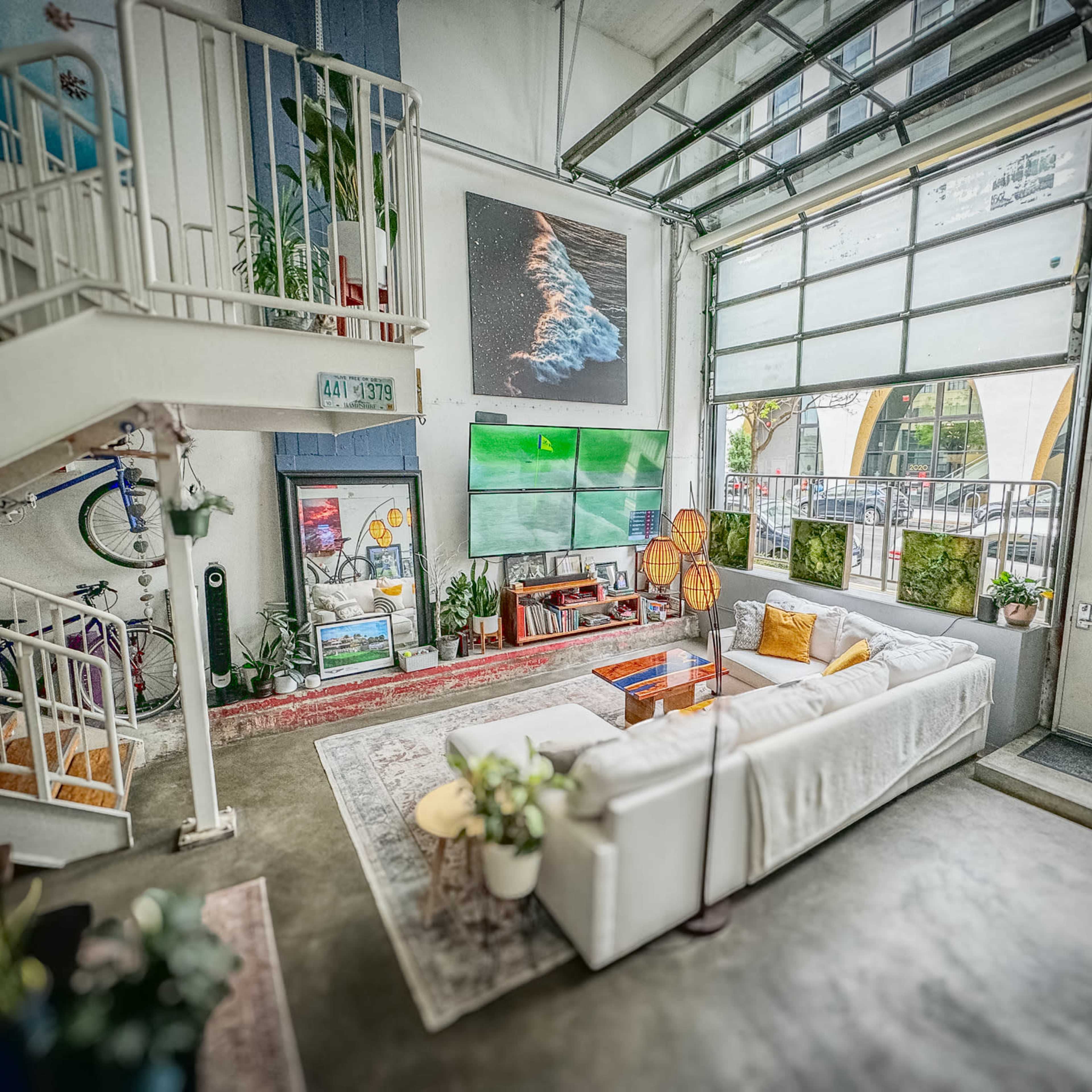 A spacious living area features a large white sofa, colorful artwork on the walls, and an open layout with large windows and greenery.