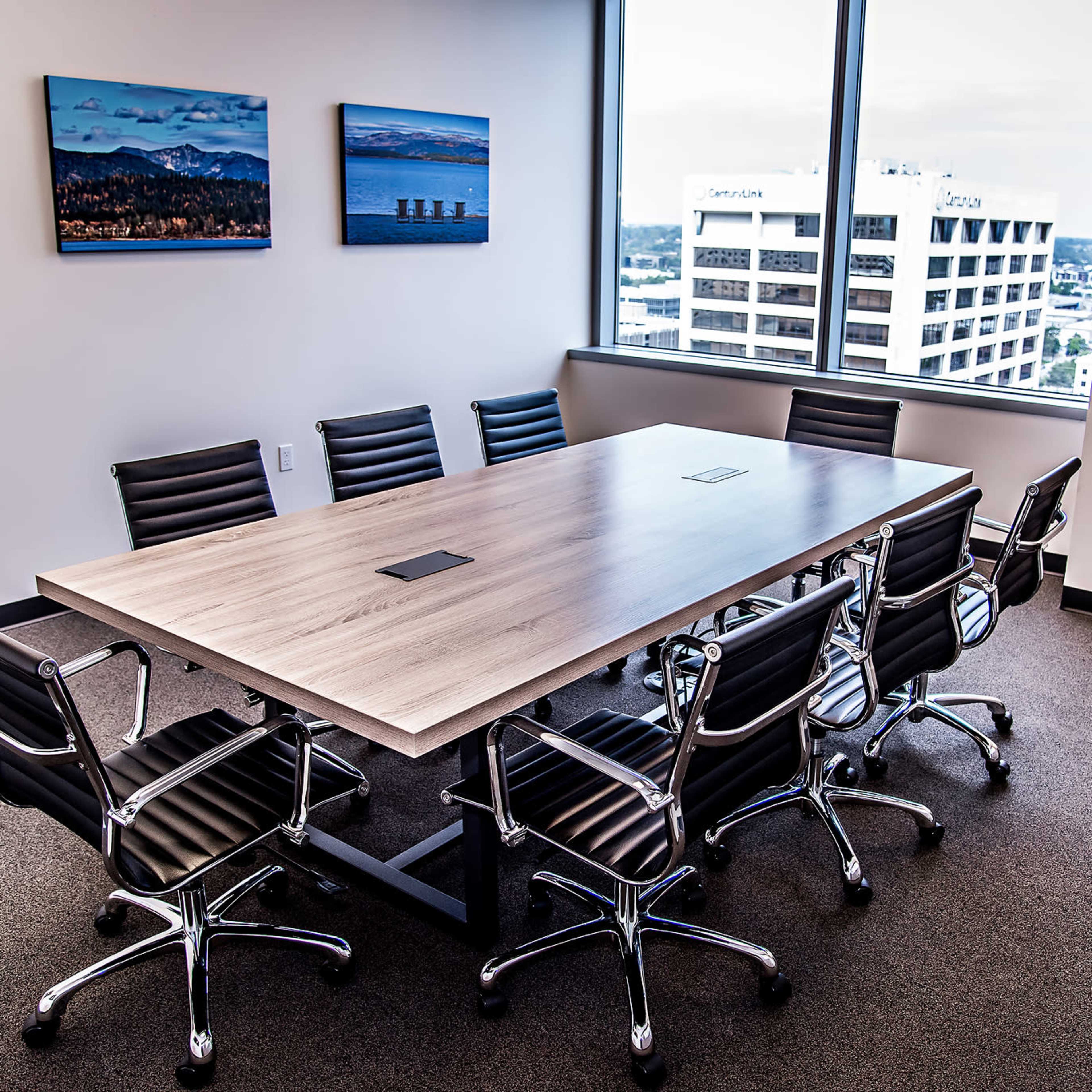 A large wooden conference table is surrounded by eight black office chairs in a well-lit meeting room featuring large windows.