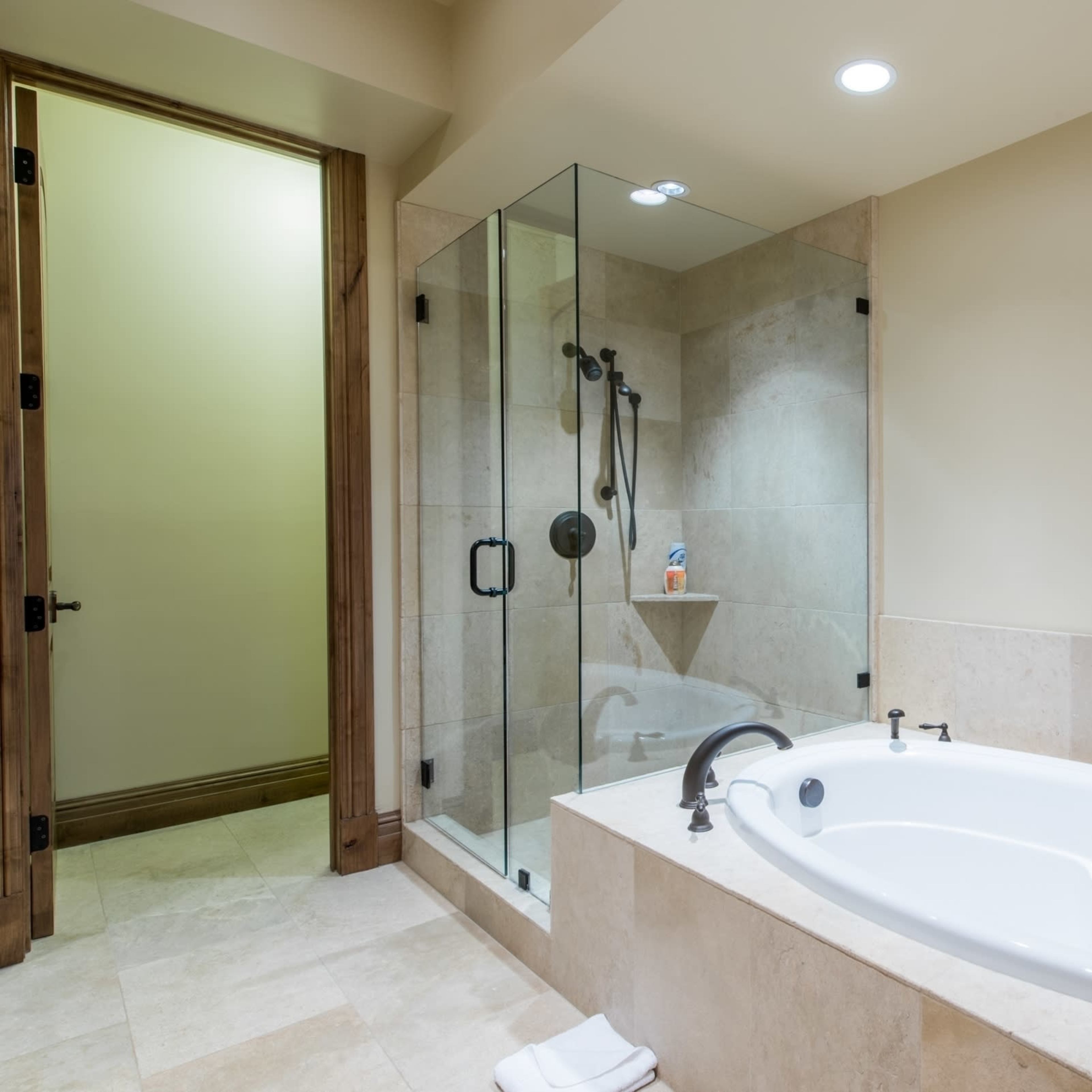 A bathroom with a glass-enclosed shower, a white bathtub, and beige stone tiles on the floor and walls.